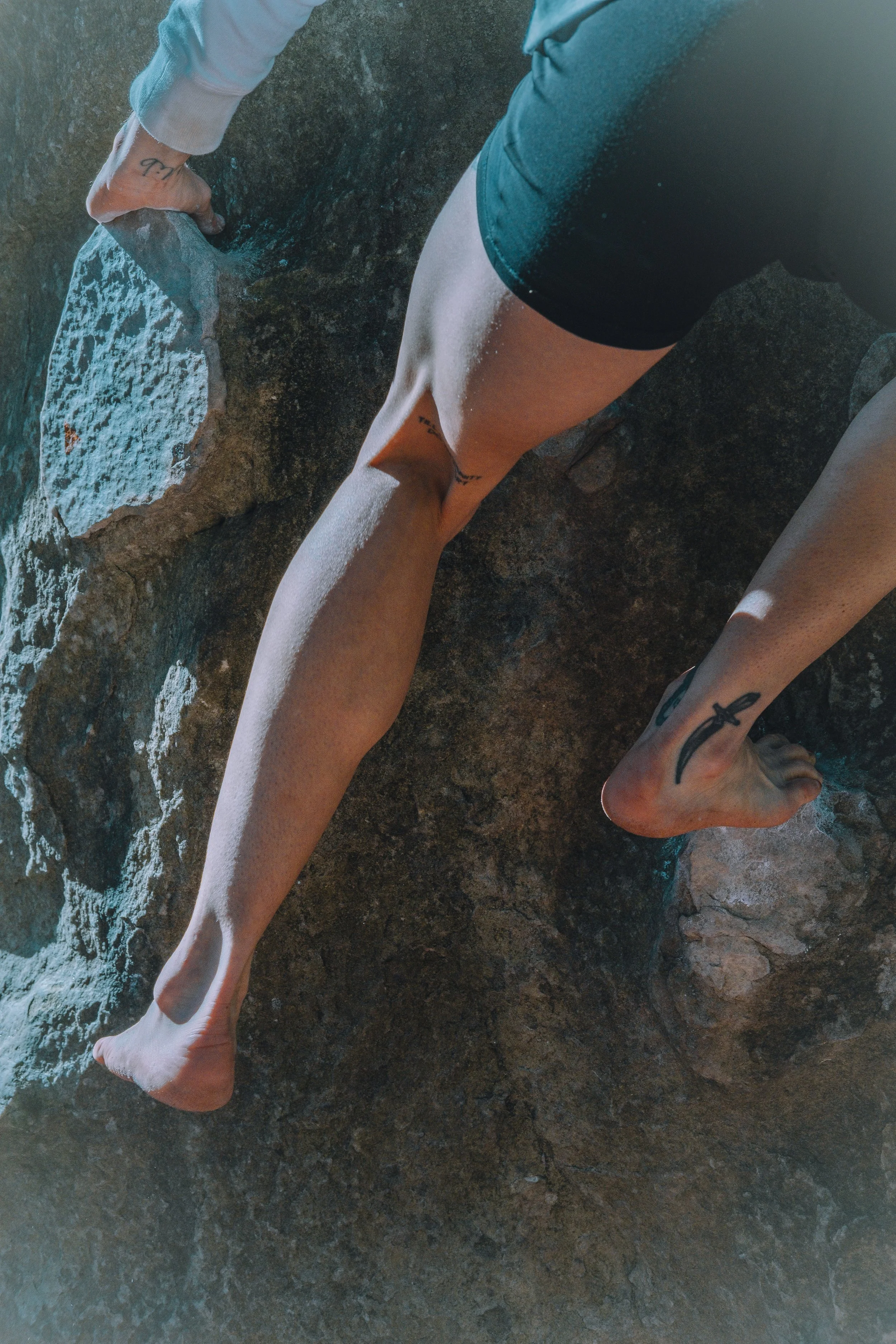 Une personne assise ou agenouillée sur une surface rocheuse, portant un short et ayant plusieurs tatouages, dont un coquillage sur la cheville. La personne ne porte pas de chaussures.