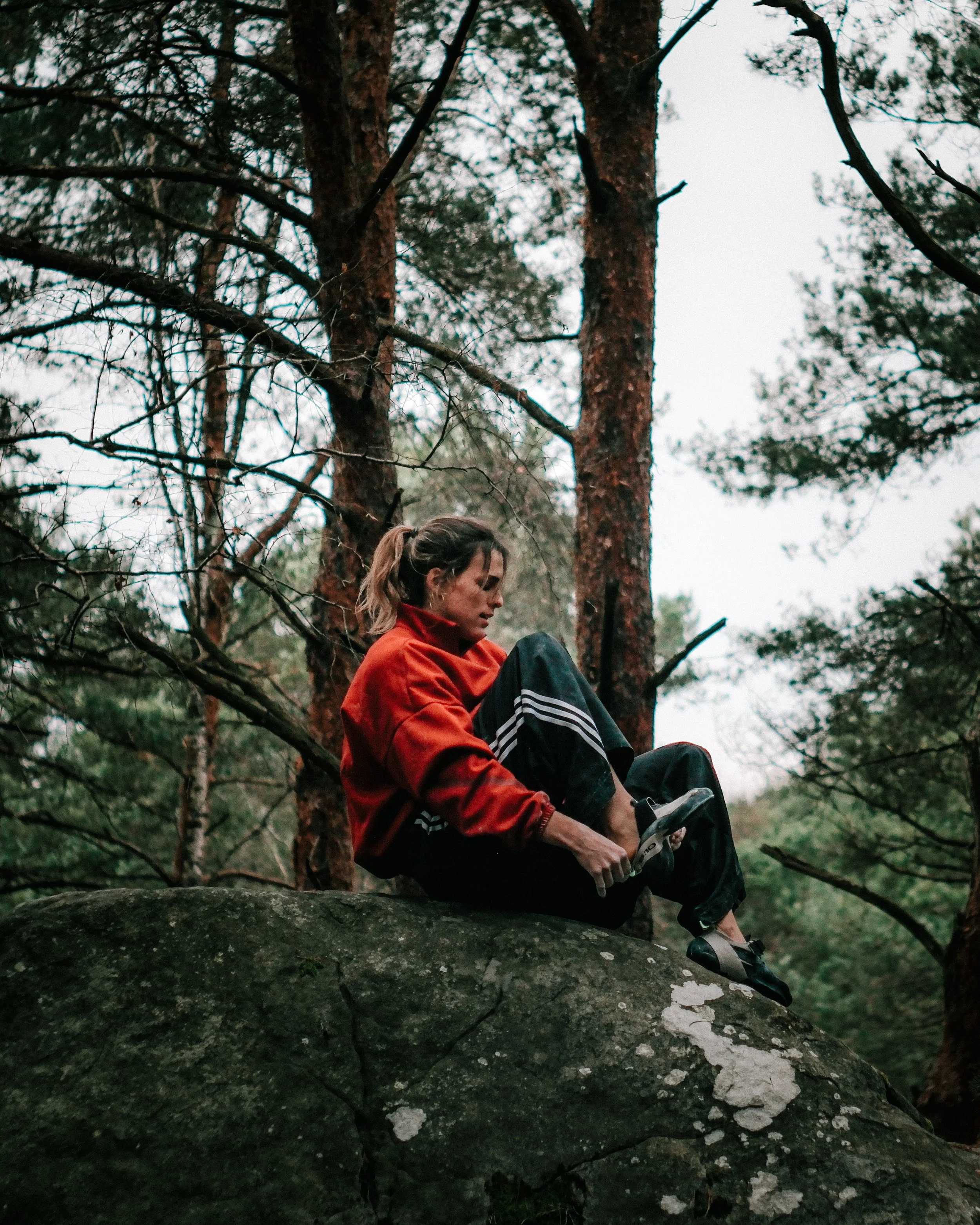 Une femme assise sur une branche d'arbre en forêt, portant une veste rouge, écouteur, avec des branches d'arbres et le ciel en arrière-plan.