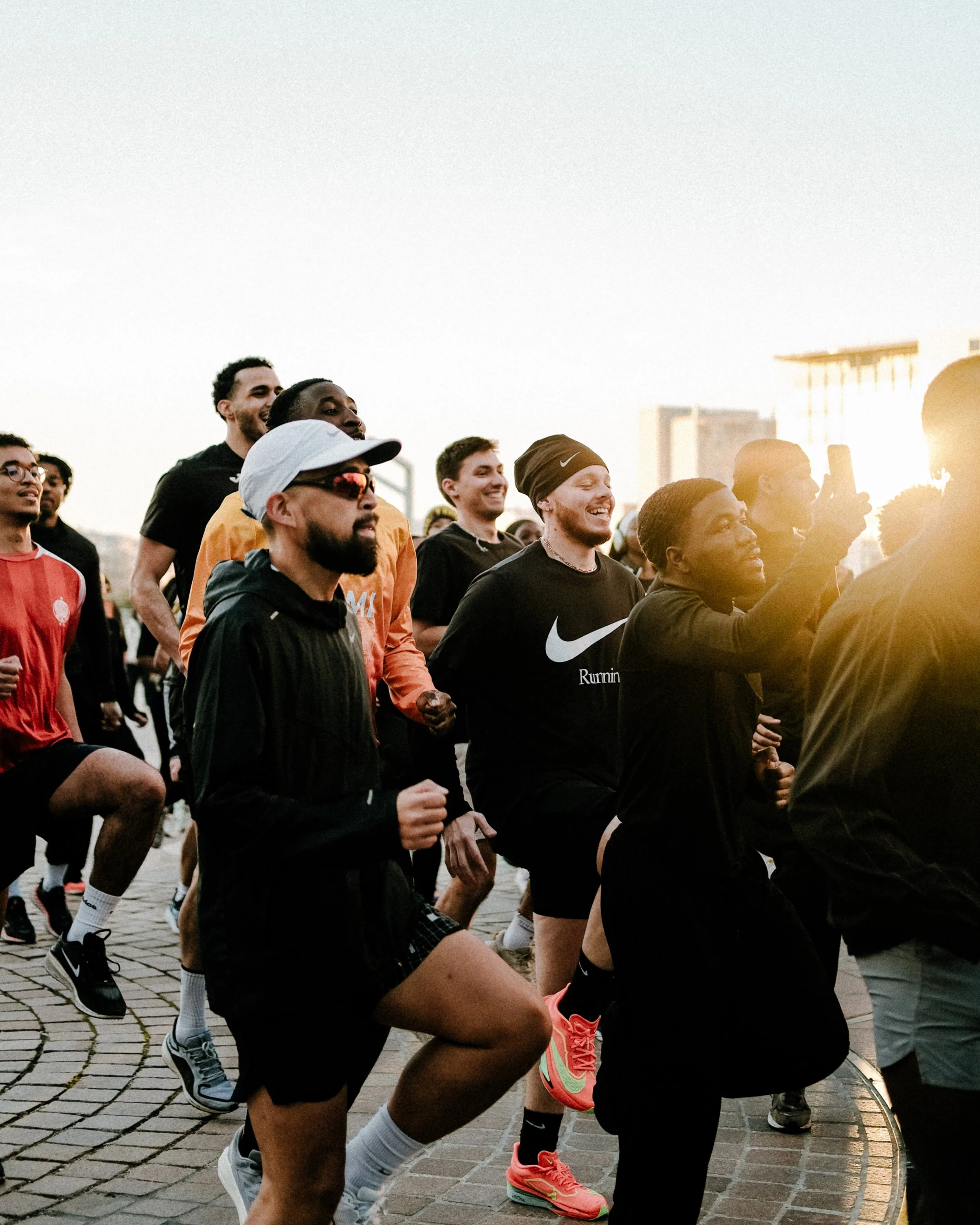 Groupe de coureurs en tenue de sport, certains souriants, en train de courir ou de s'échauffer dans une place pavée, avec des bâtiments en arrière-plan, au coucher du soleil.