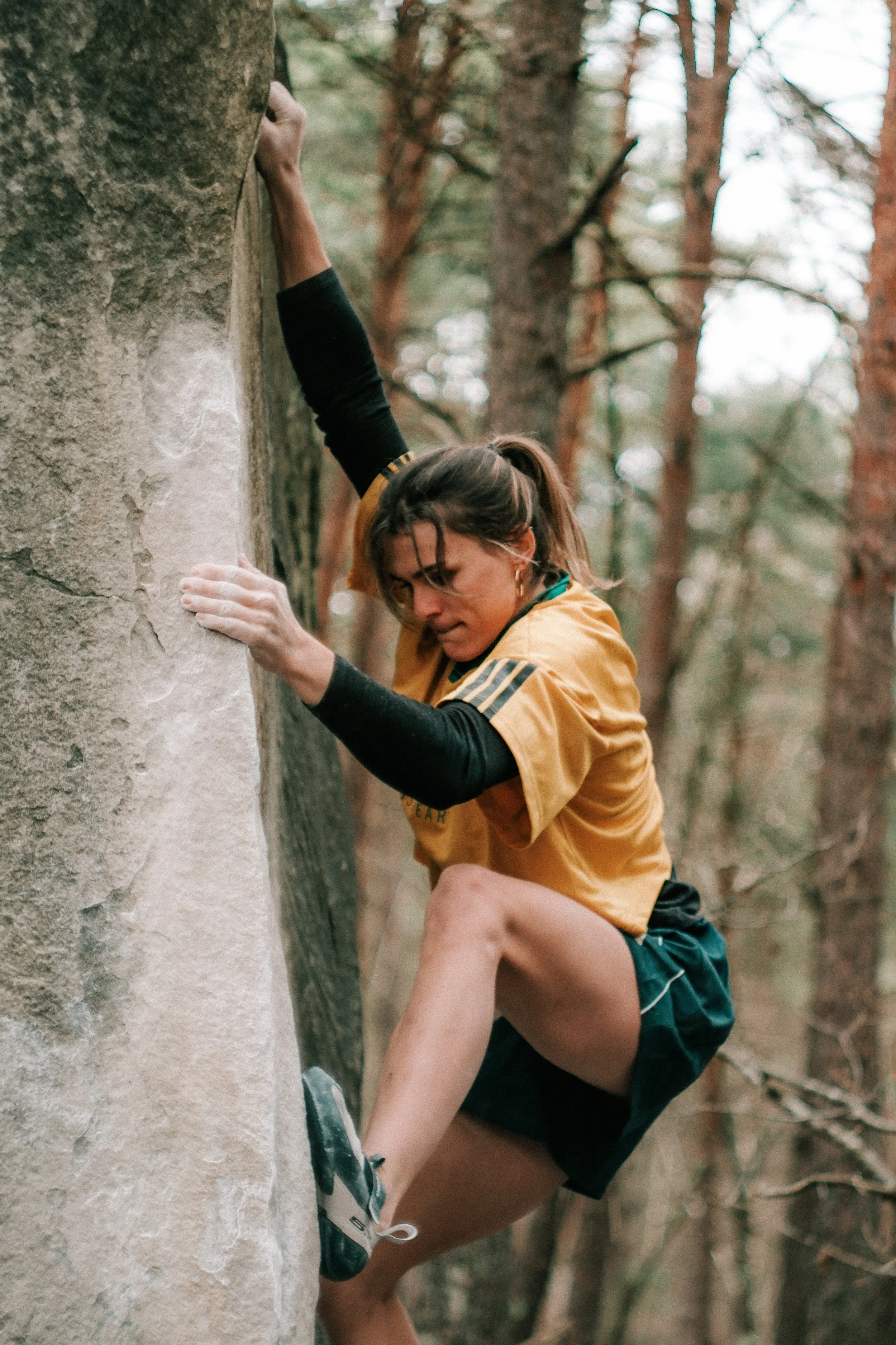 Femme faisant de l'escalade en extérieur dans une forêt, portant un t-shirt orange et un short noir.