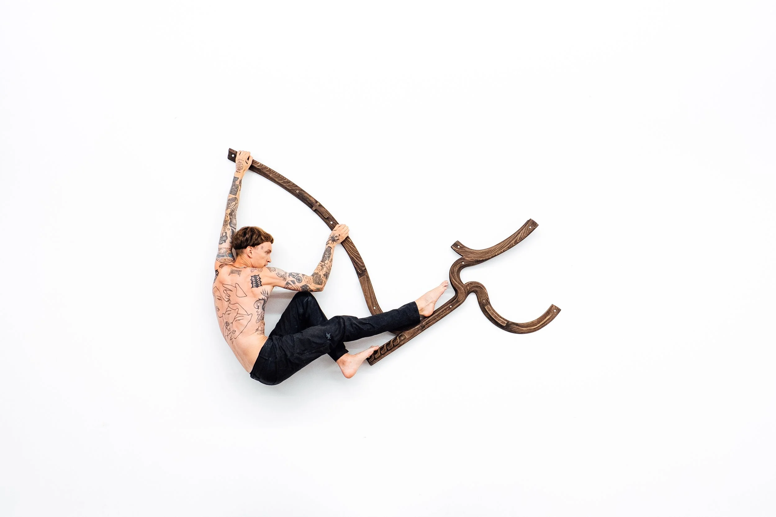 Un jeune homme nu, tatoué, assis sur une sculpture en bois, dans un espace blanc.