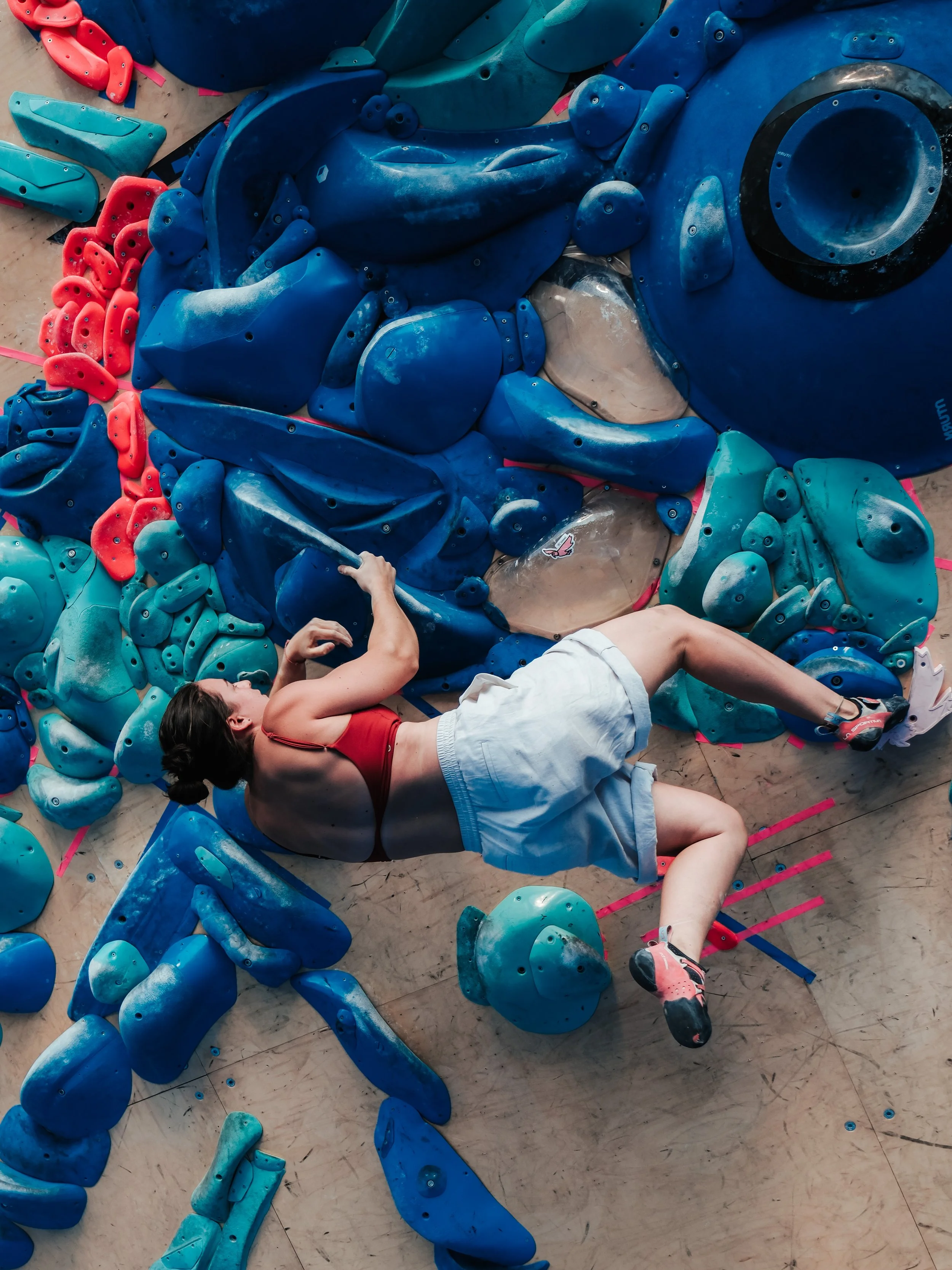 Une femme allongée sur un tapis d'escalade intérieure, entourée de prises de couleurs bleue, turquoise et rouge.