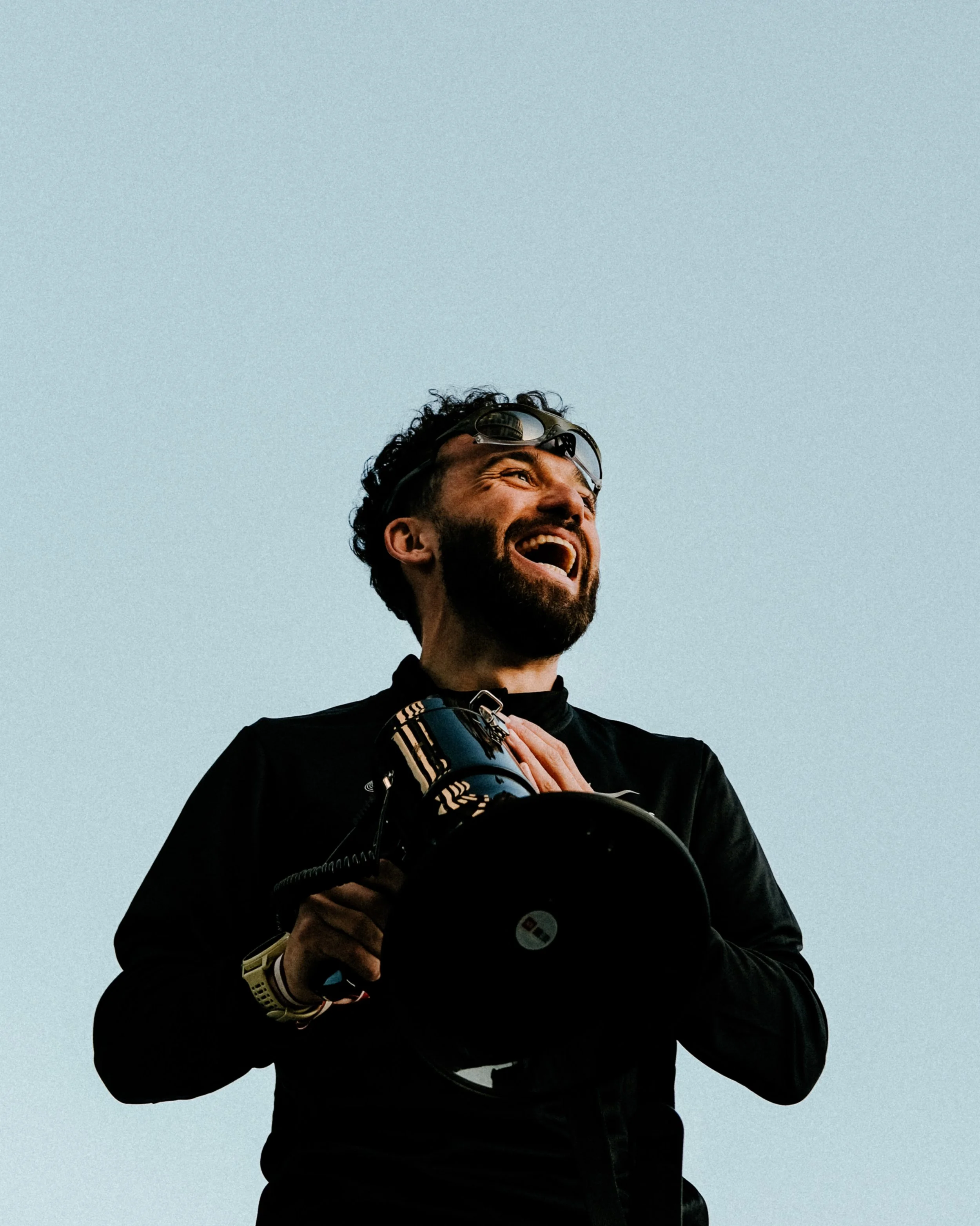 Homme souriant avec des lunettes de soleil sur la tête et un suivi d'un appareil photo dans une main, portant une montre, contre un ciel clair.