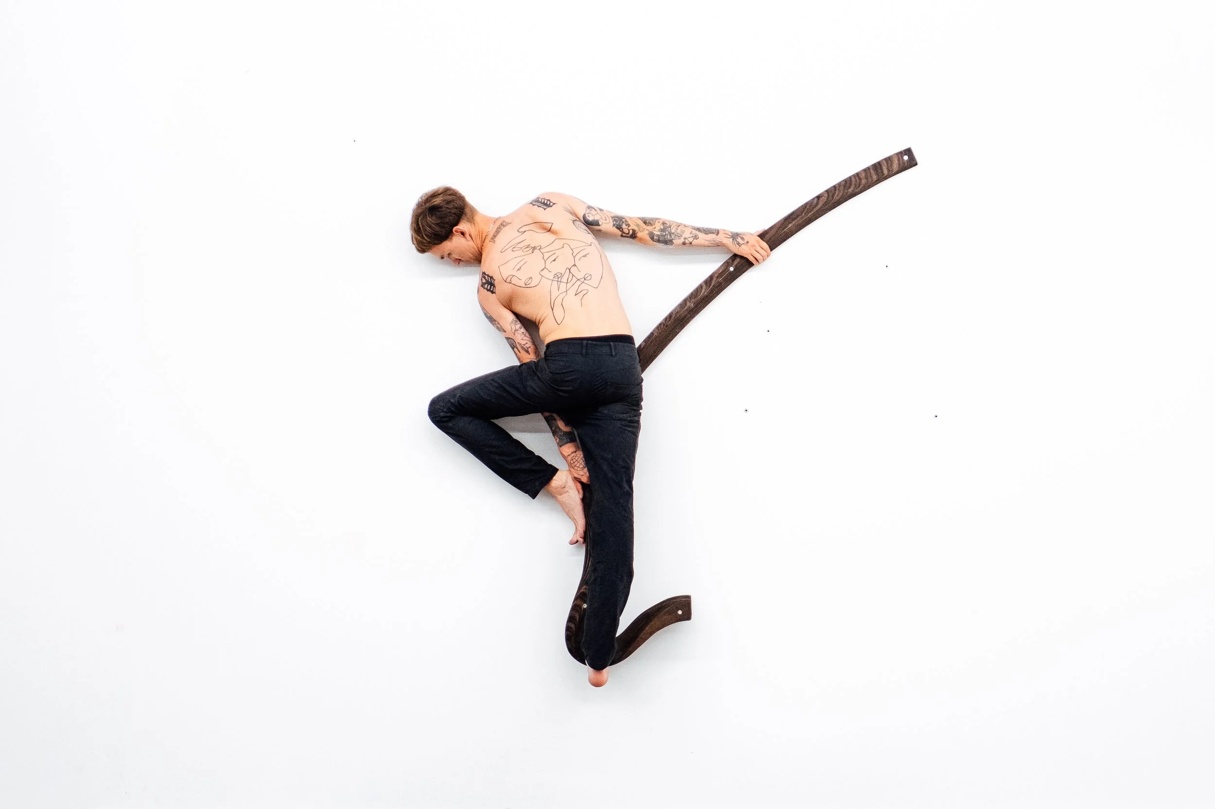 Un homme nu tatoué en position de yoga sur un mur blanc, utilisant une rampe en bois plate.