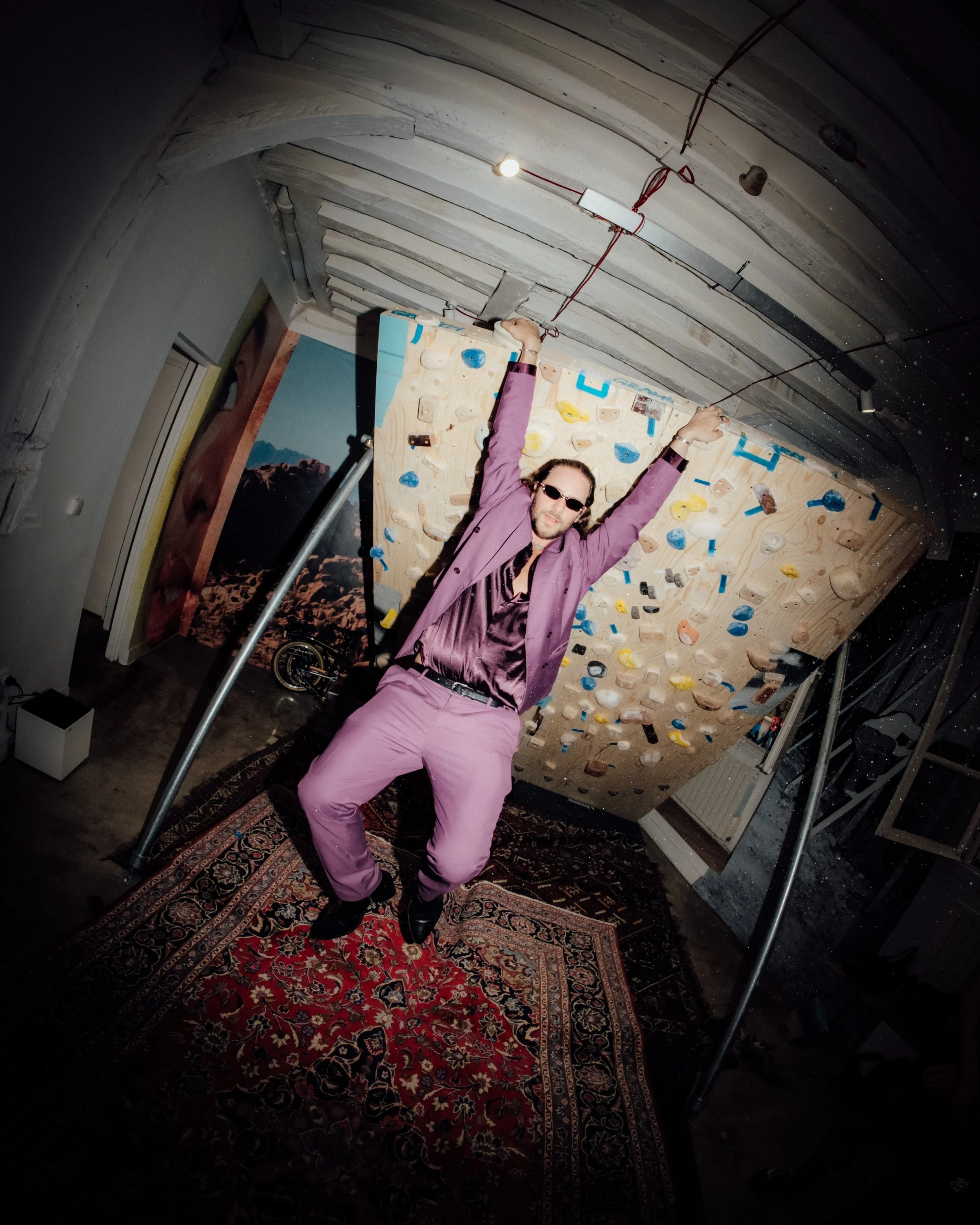 Une personne portant des lunettes de soleil, un costume violet, et avec des cheveux longs, fait une pose dynamique en levant ses bras, debout devant un mur d'escalade intérieur. La pièce est décorée avec un tapis persan sur le sol, un mur peint avec 