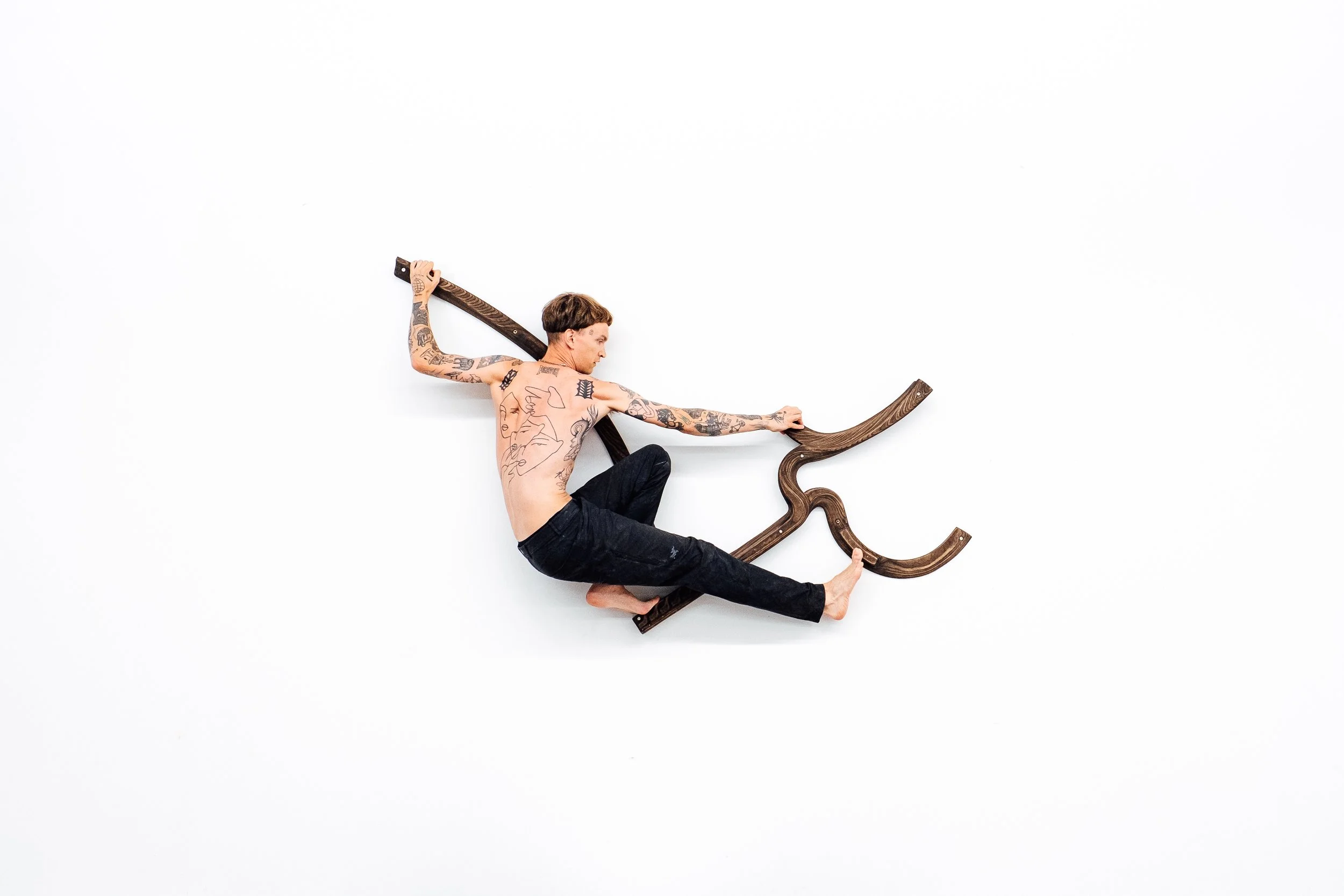 Un homme tatoué sans vêtement, assis sur un objet en bois qui ressemble à une pièce de mobilier, dans une pièce avec un mur blanc, tenant une planche en bois.