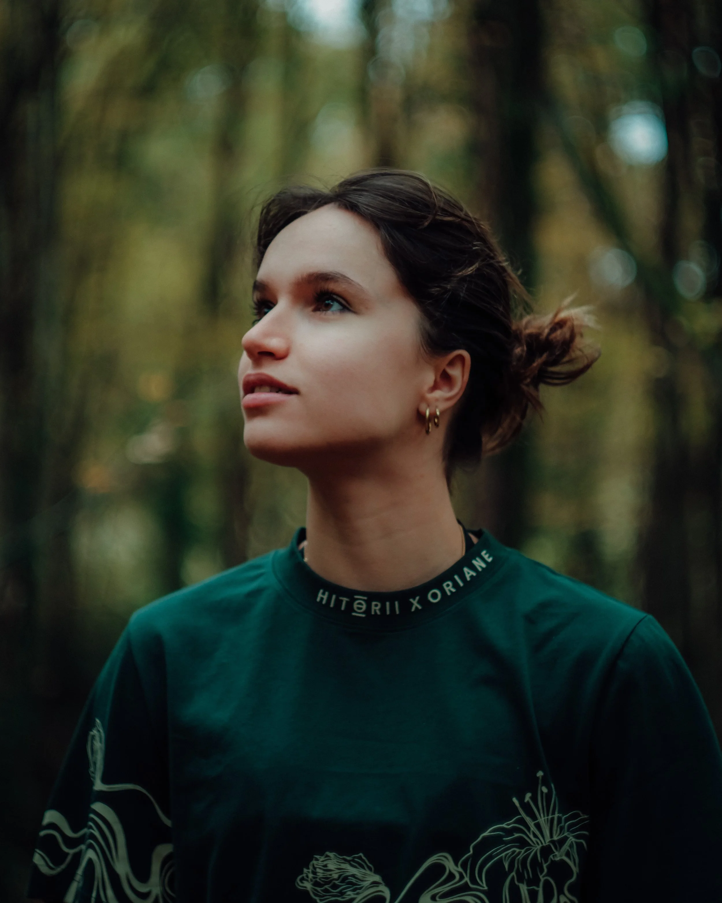 Portrait d'une jeune femme aux cheveux bruns courts et bouclés, portant un haut vert foncé avec un col rond, se tenant dans un environnement naturel avec des arbres flous en arrière-plan.