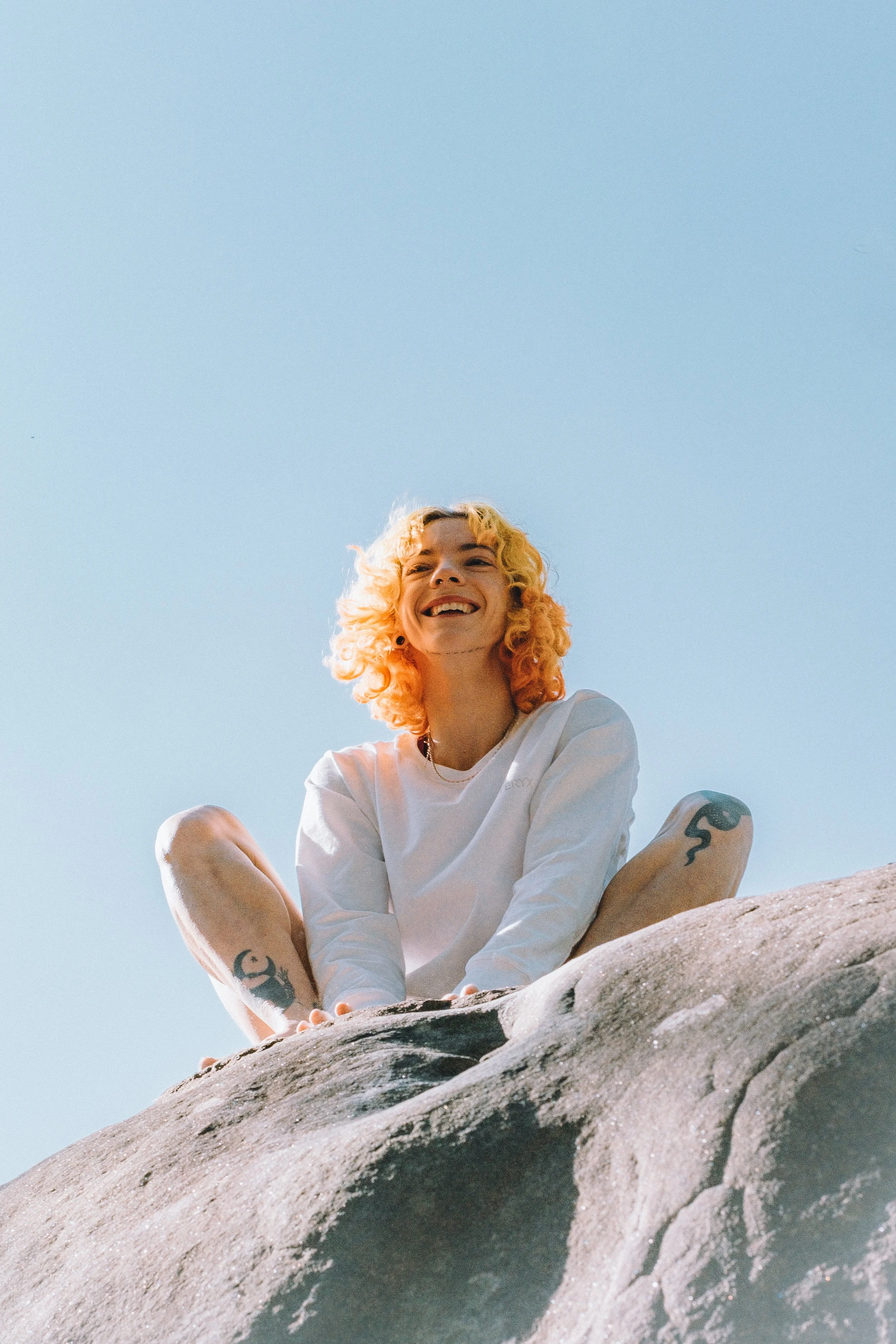 Jeune femme aux cheveux blonds bouclés assise sur une roche, souriante, contre un ciel bleu clair.
