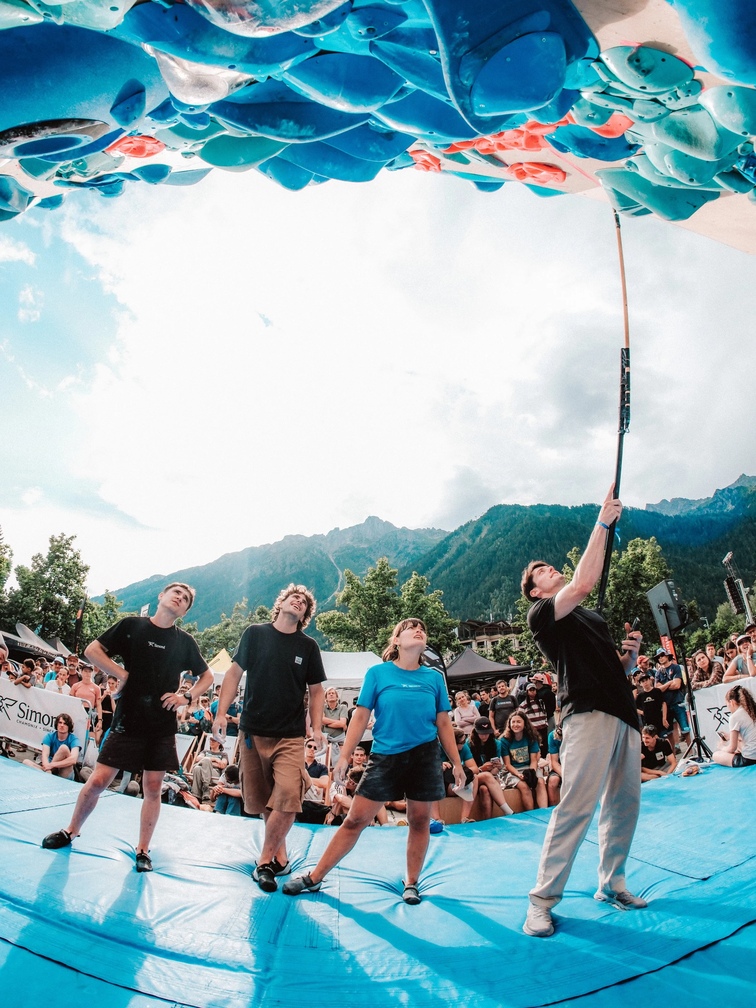 Un groupe de personnes, dont certains portent des T-shirts noirs et un vêtement bleu, participent à un événement de relay, en train de tirer sur une corde dans un environnement extérieur avec une foule, des tentes, des montagnes en arrière-plan et un