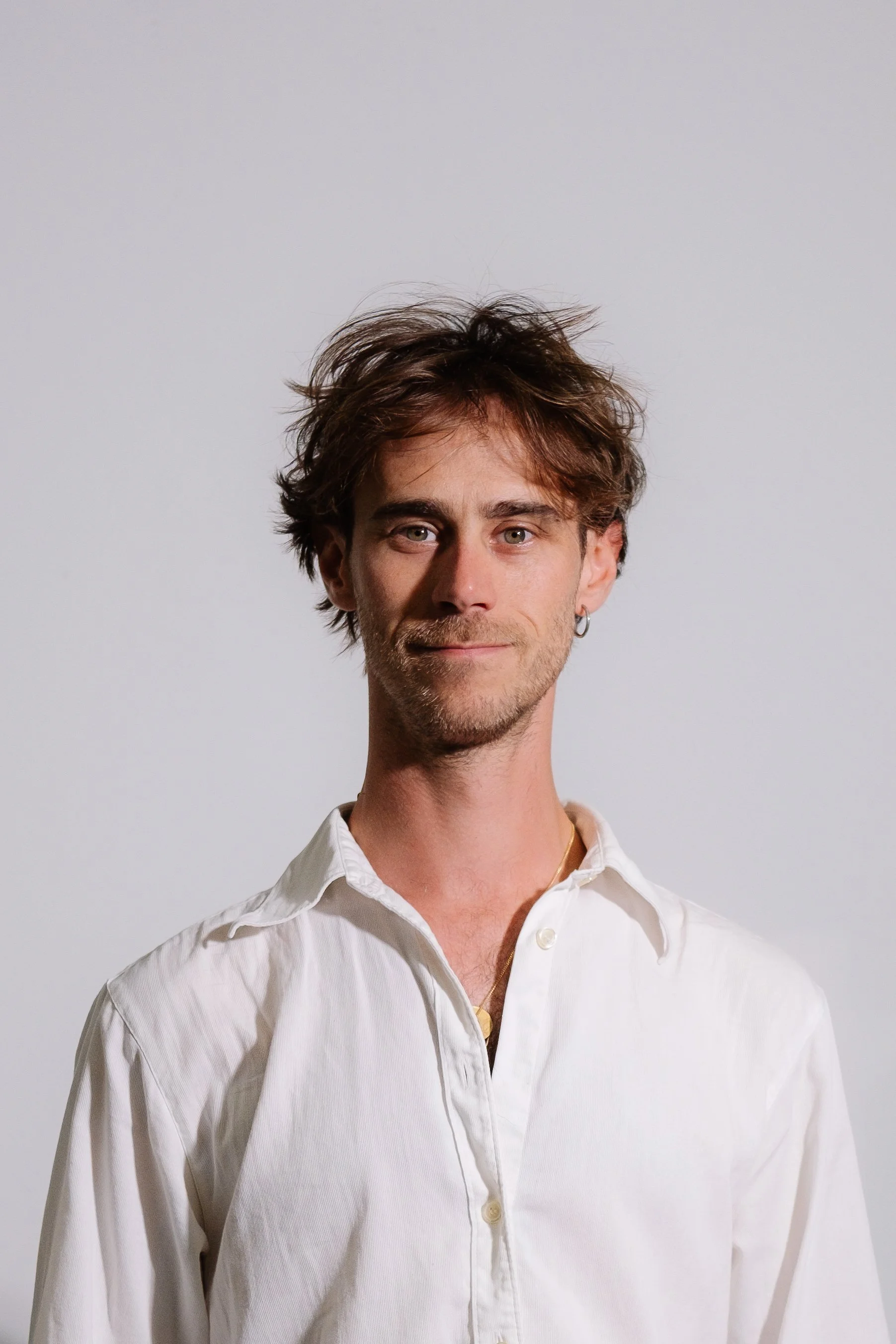 Portrait d'un homme avec des cheveux bruns en désordre, portant une chemise blanche ouverte, souriant légèrement, contre un fond blanc.