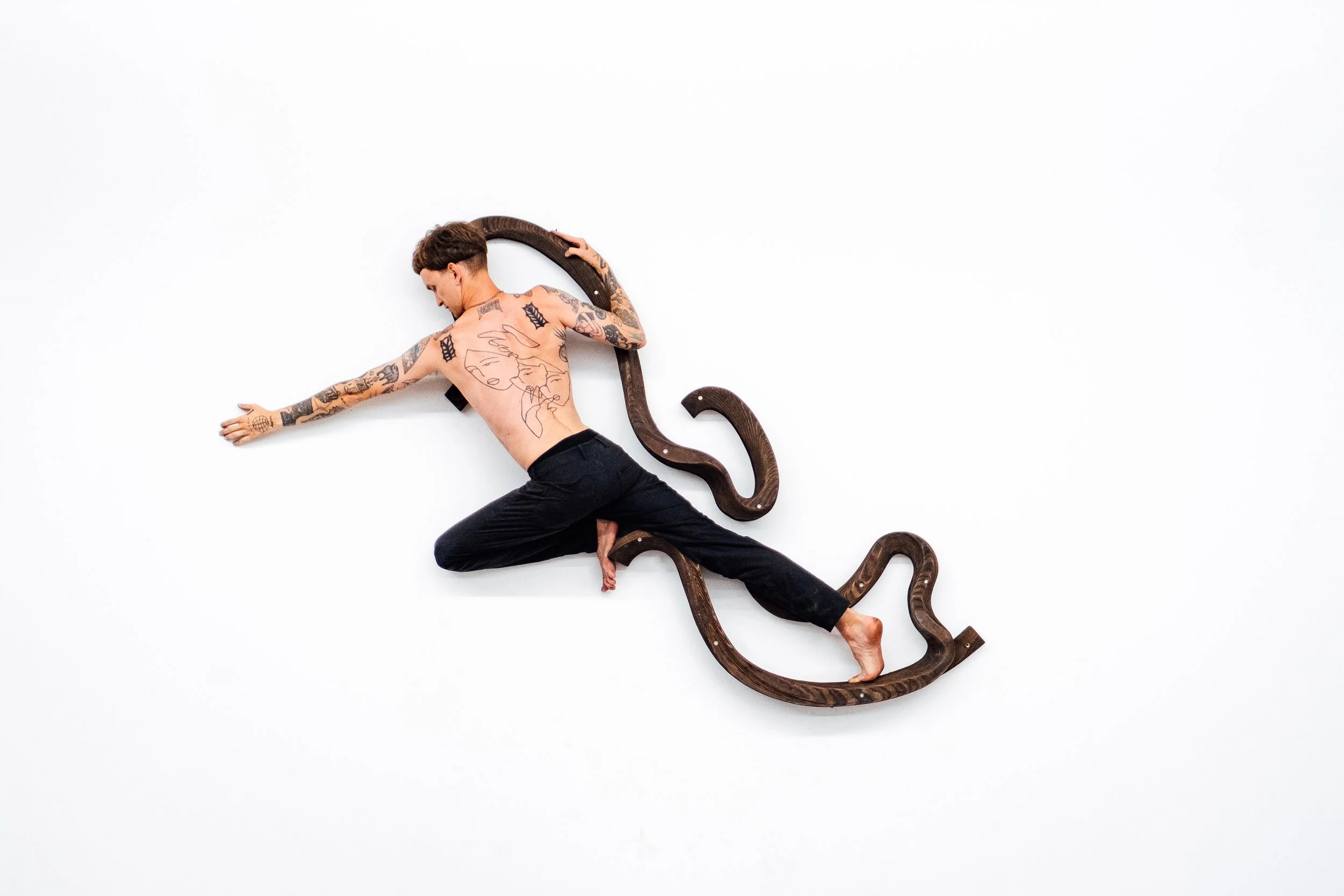Une personne nue tatouée, allongée sur un fond blanc, entourée de serpents en bois, dans une pose artistique.