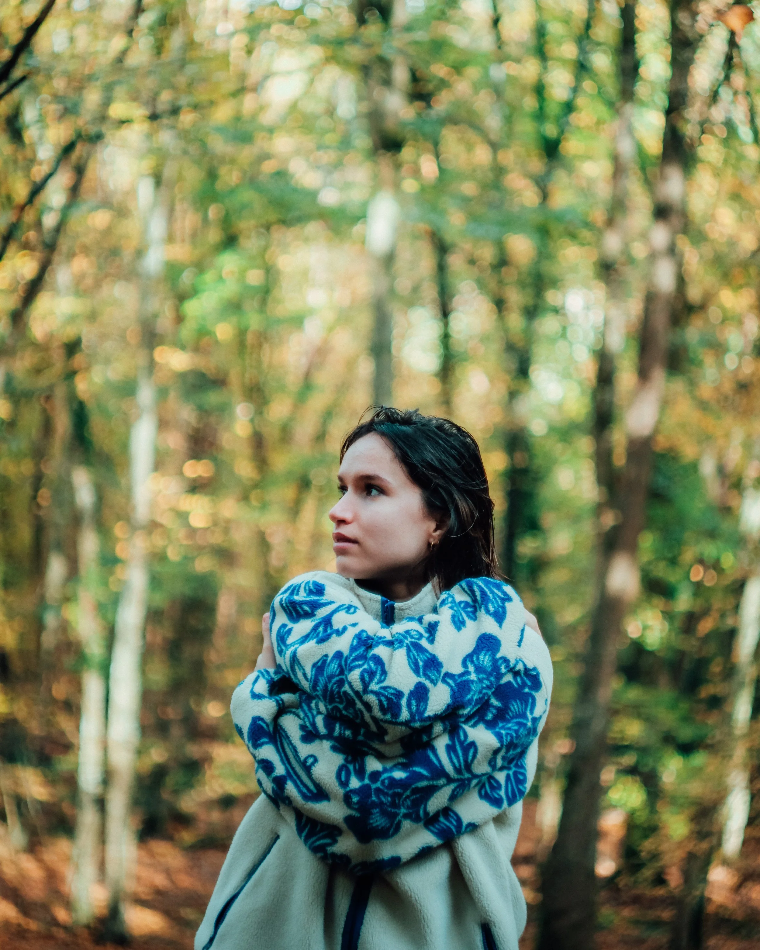 Une femme avec des cheveux noirs, portant un pull à motifs bleus et blancs, se tient dans une forêt d'automne avec des feuilles orangées et jaunes, regardant légèrement sur le côté.