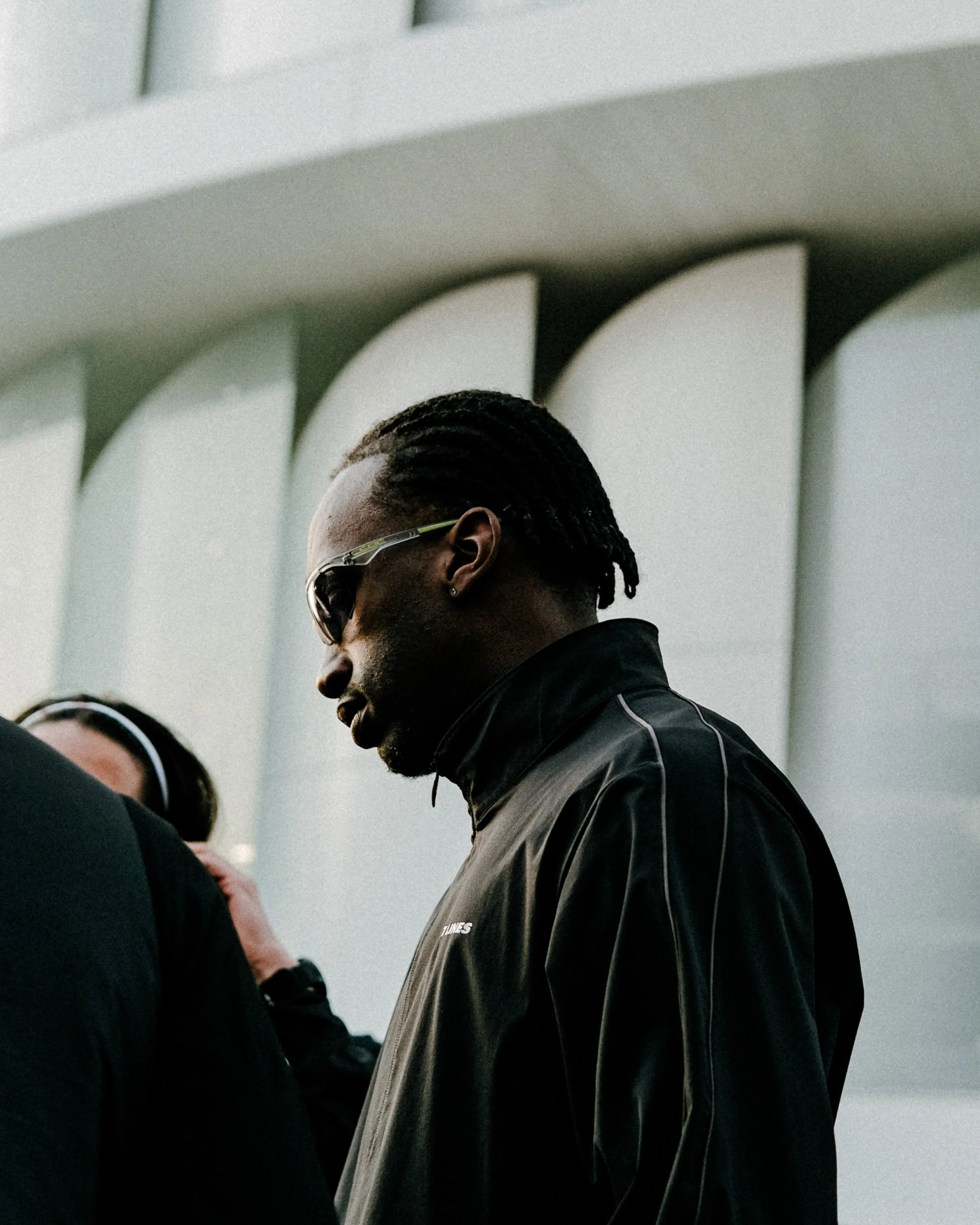 Un homme portant des lunettes de soleil noires et un blouson noir est photographié en profil, dans un environnement intérieur. Une personne portant un uniforme ou une veste avec une inscription partiellement visible 'TUNES' est également présente.