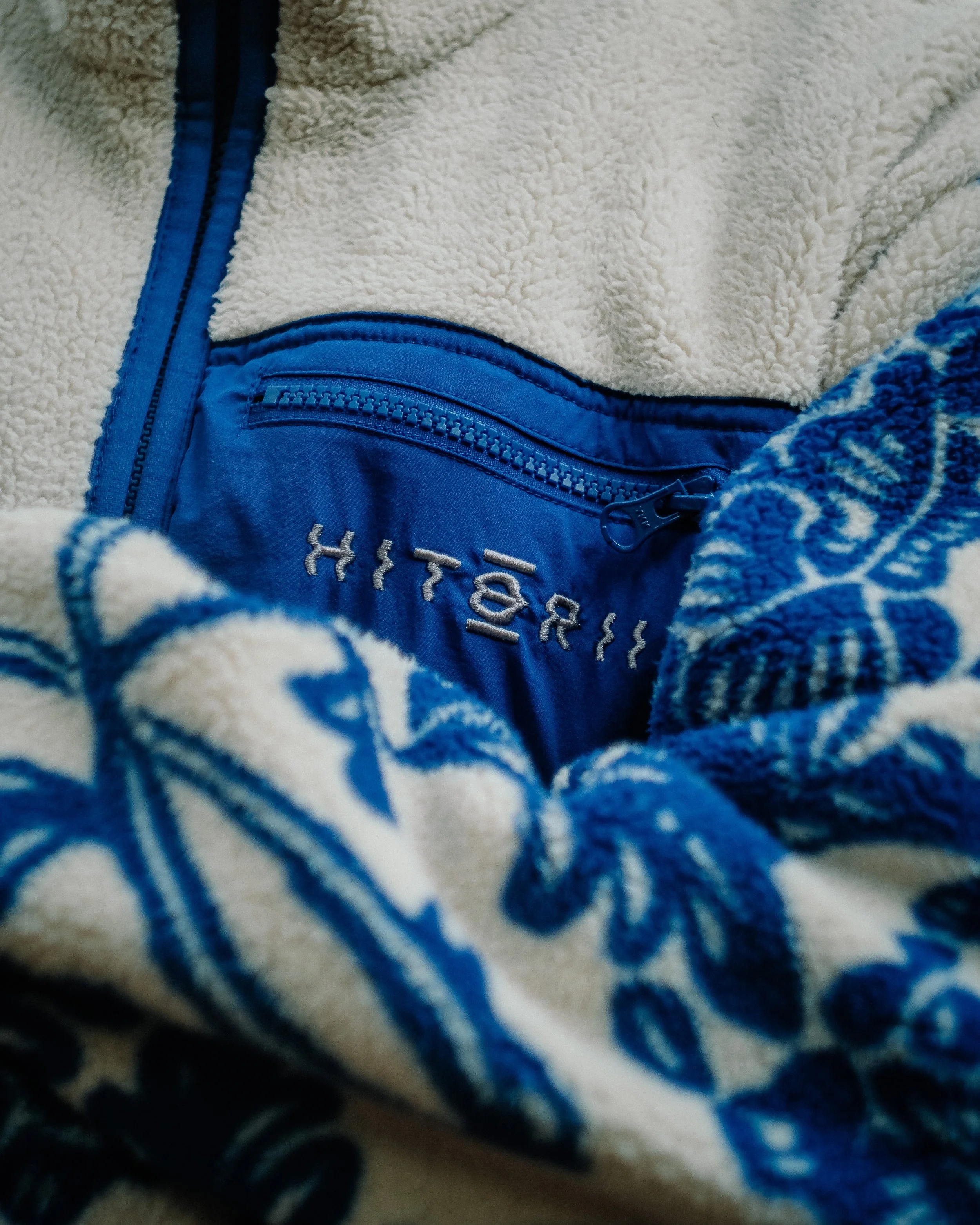 Photo d’une fermeture éclair bleue sur un vêtement en tissu sherpa beige, avec une partie en tissu à motif bleu et blanc visible en bas.