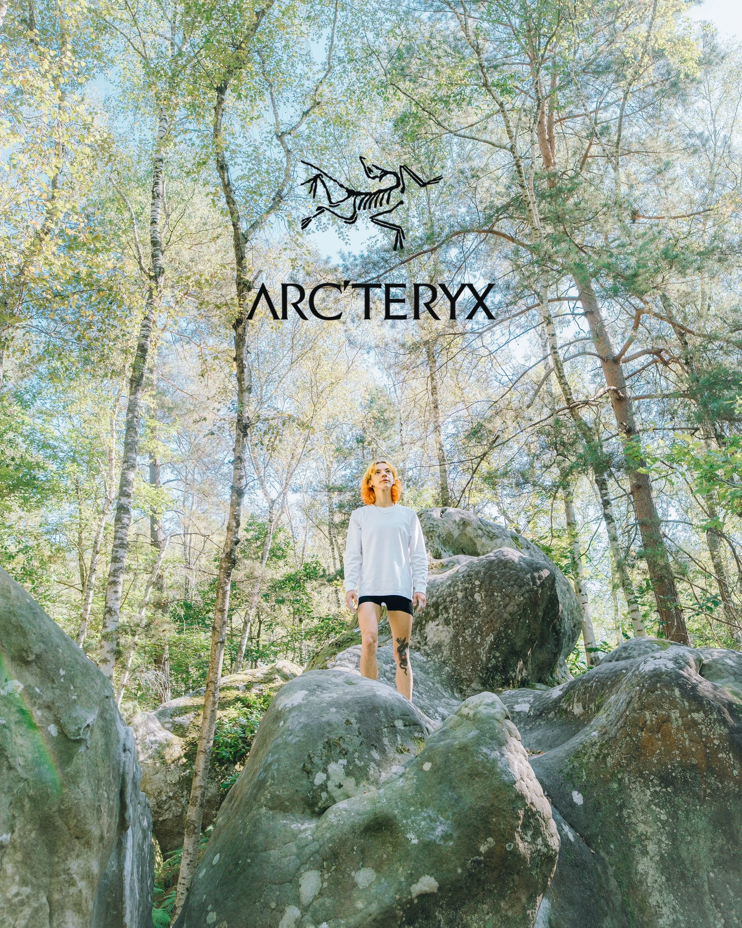 Une femme avec les cheveux roux et une tattoo sur la jambe, vêtue d'un sweat blanc et d'un short noir, se tient debout sur des rochers dans une forêt verdoyante avec des arbres et un ciel bleu en arrière-plan.