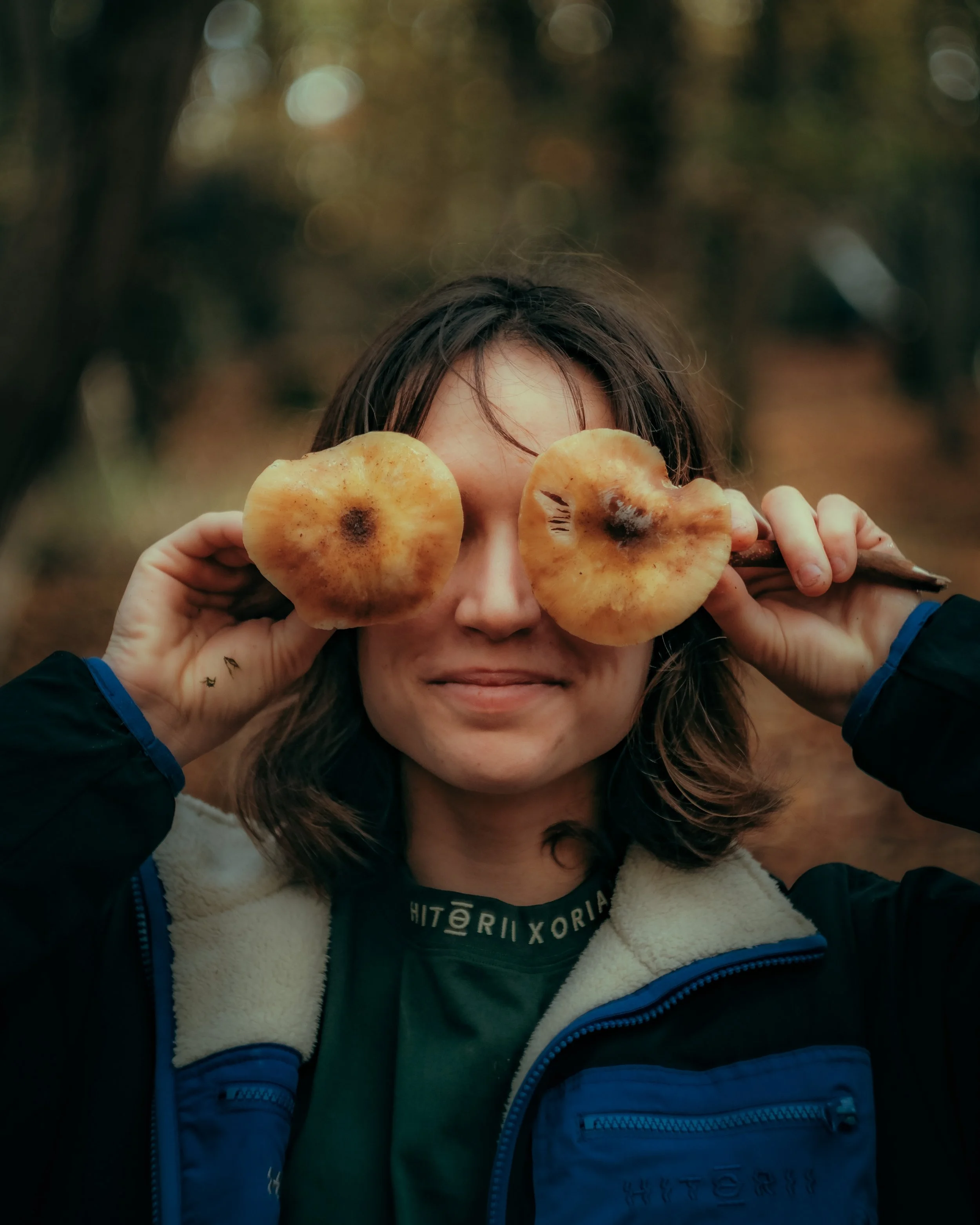 Jeune fille avec des cheveux bruns, portant une veste noire à doublure en peluche beige et une écharpe, tenant deux champignons devant ses yeux dans un décor naturel d'automne, flou à l'arrière-plan.