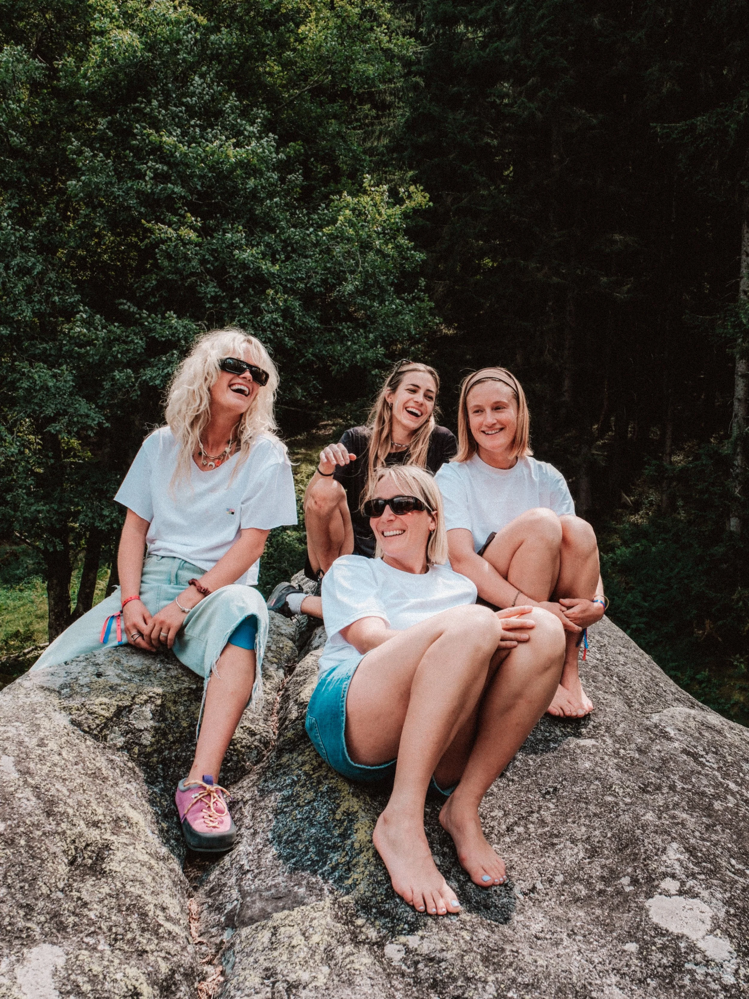 Groupe de cinq femmes souriantes et détendues, assises et allongées sur des rochers en pleine nature, entourées d'arbres verts.