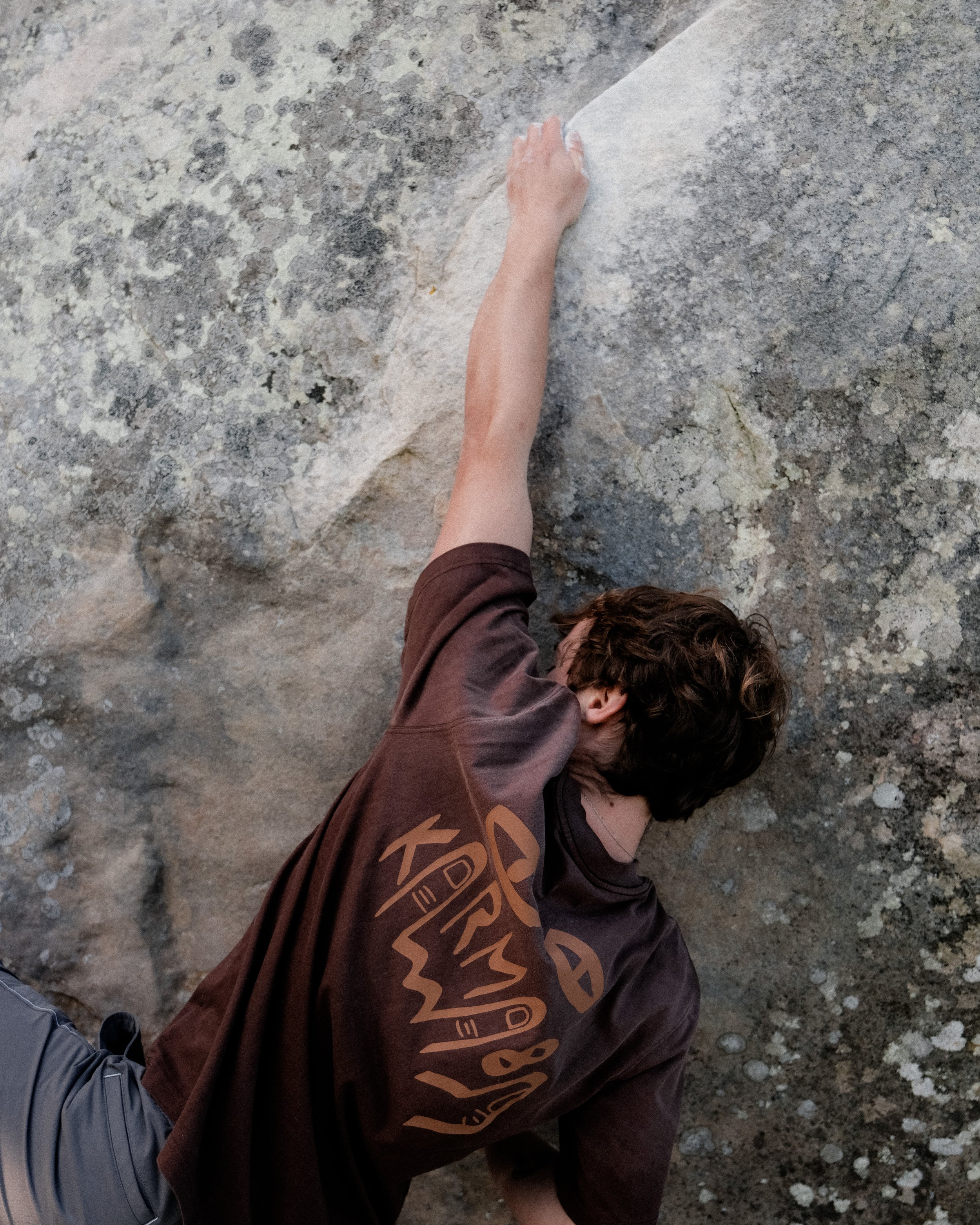 Une personne grimpe une paroi rocheuse en utilisant une prise. La personne porte un t-shirt marron avec un logo, un grimpeur équipé d'un sac à dos et un pantalon gris. La surface rocheuse est granuleuse avec des couleurs grises et beige. La personne 
