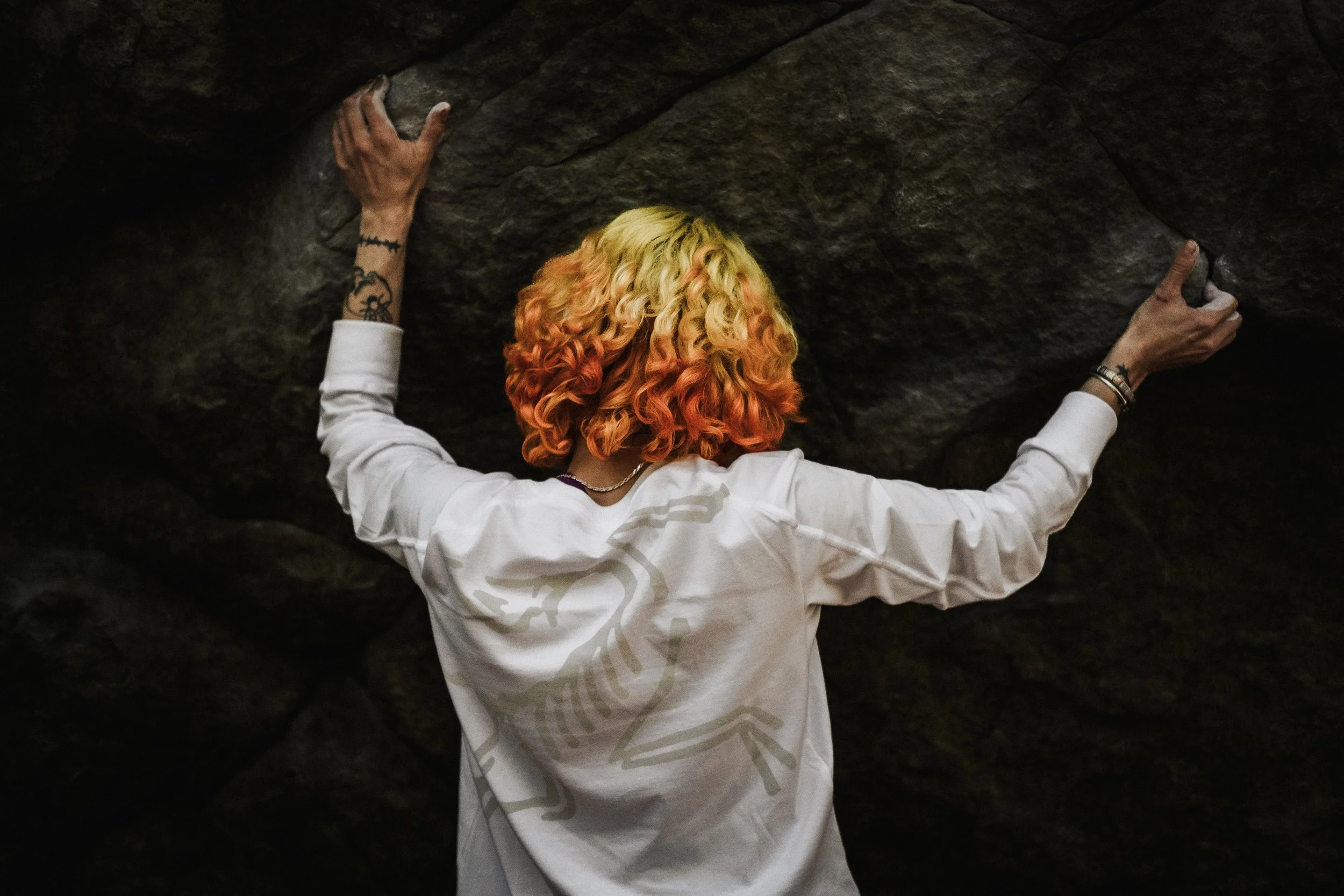 Une personne avec des cheveux bouclés teints en blond, orange et rose, qui grimpe ou explore une surface rocheuse ou un mur d'escalade, vue de dos, portant un vêtement blanc.