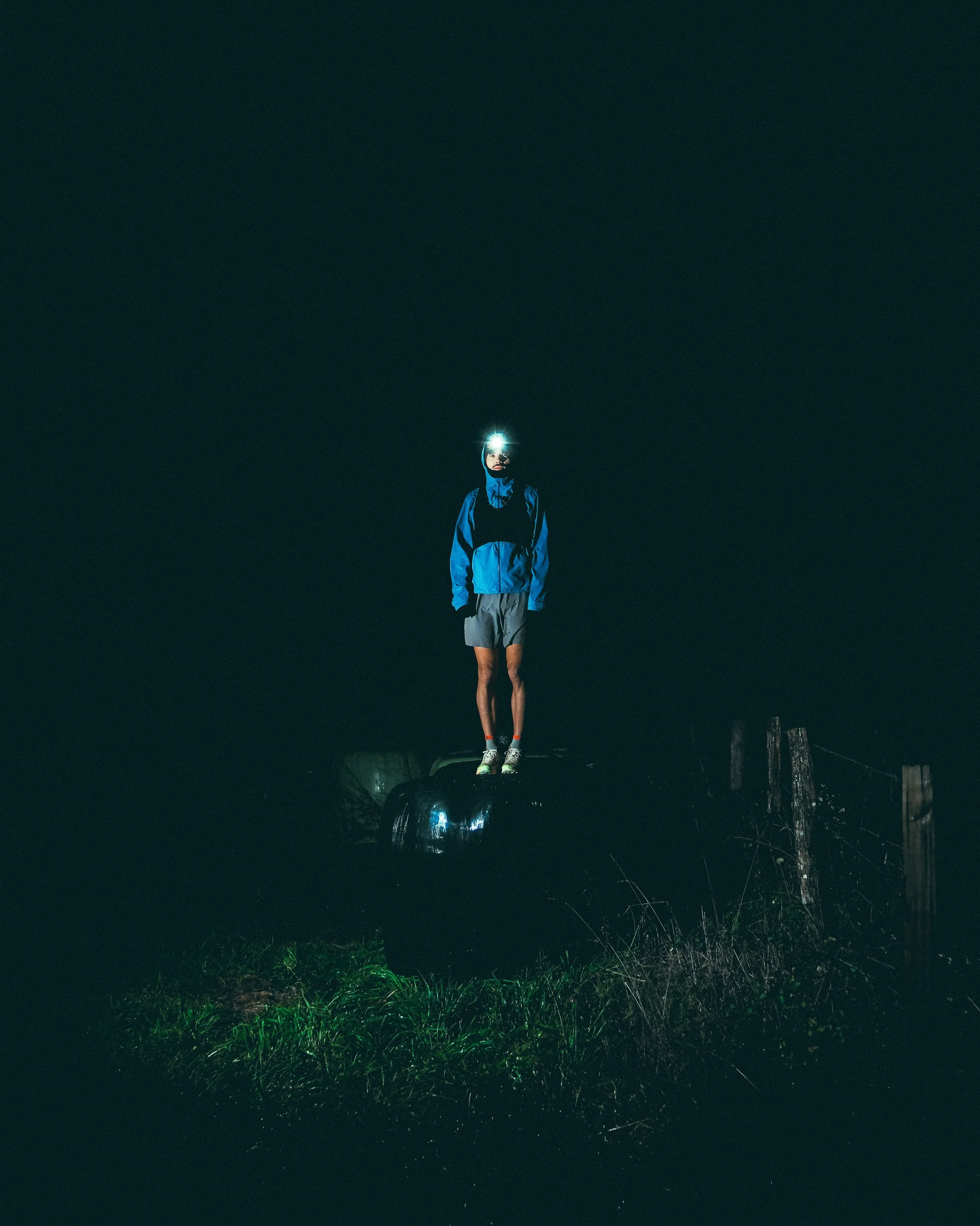 Une personne debout sur une surface noire, portant une veste et un casque avec une lampe frontale, lors d'une activité nocturne en plein air, avec de l'herbe éclairée en vert en bas de l'image.