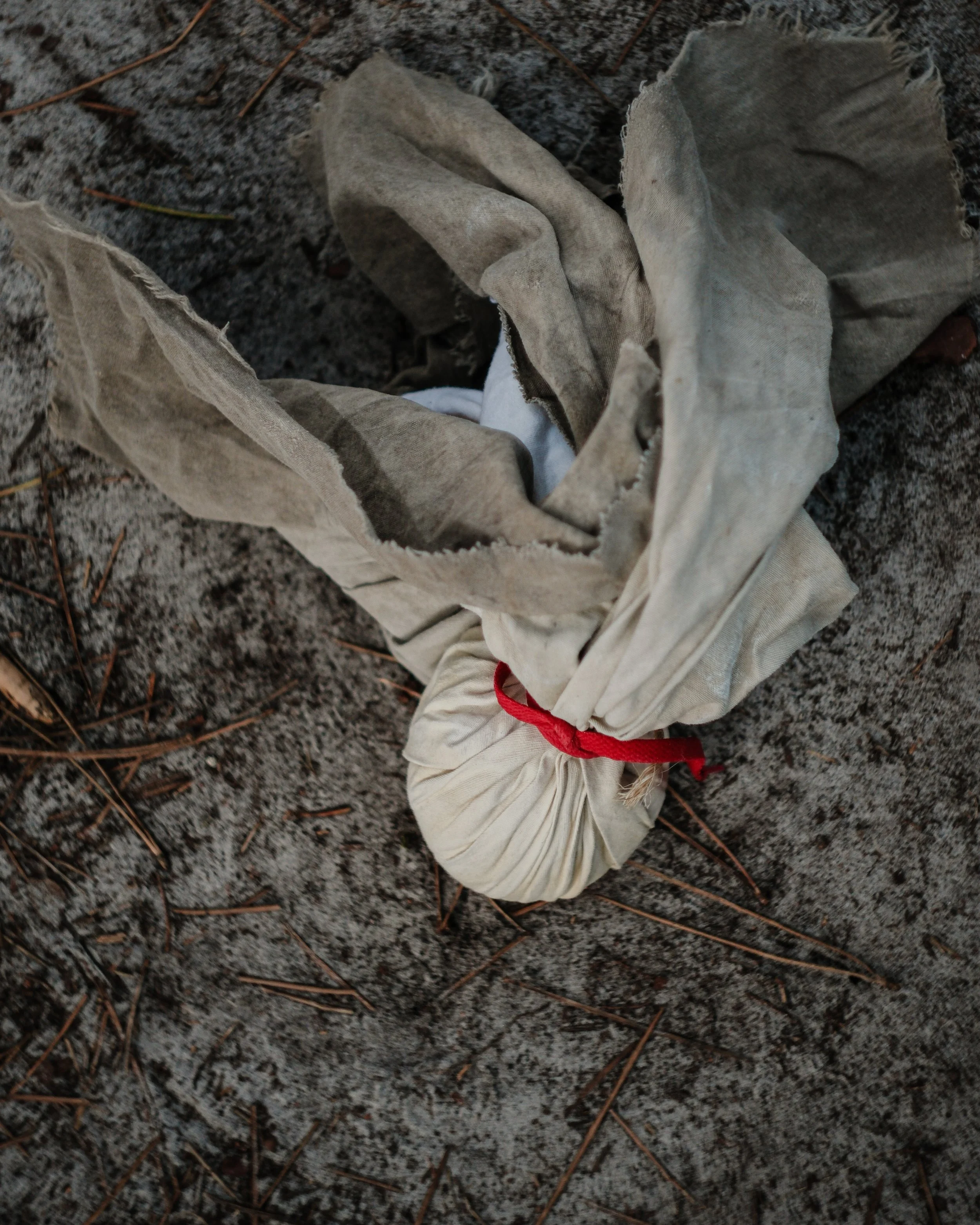 Une vieille sac en tissu blanc, sale et froissée, posée sur le sol de terre battue avec des petits cailloux et des brindilles.