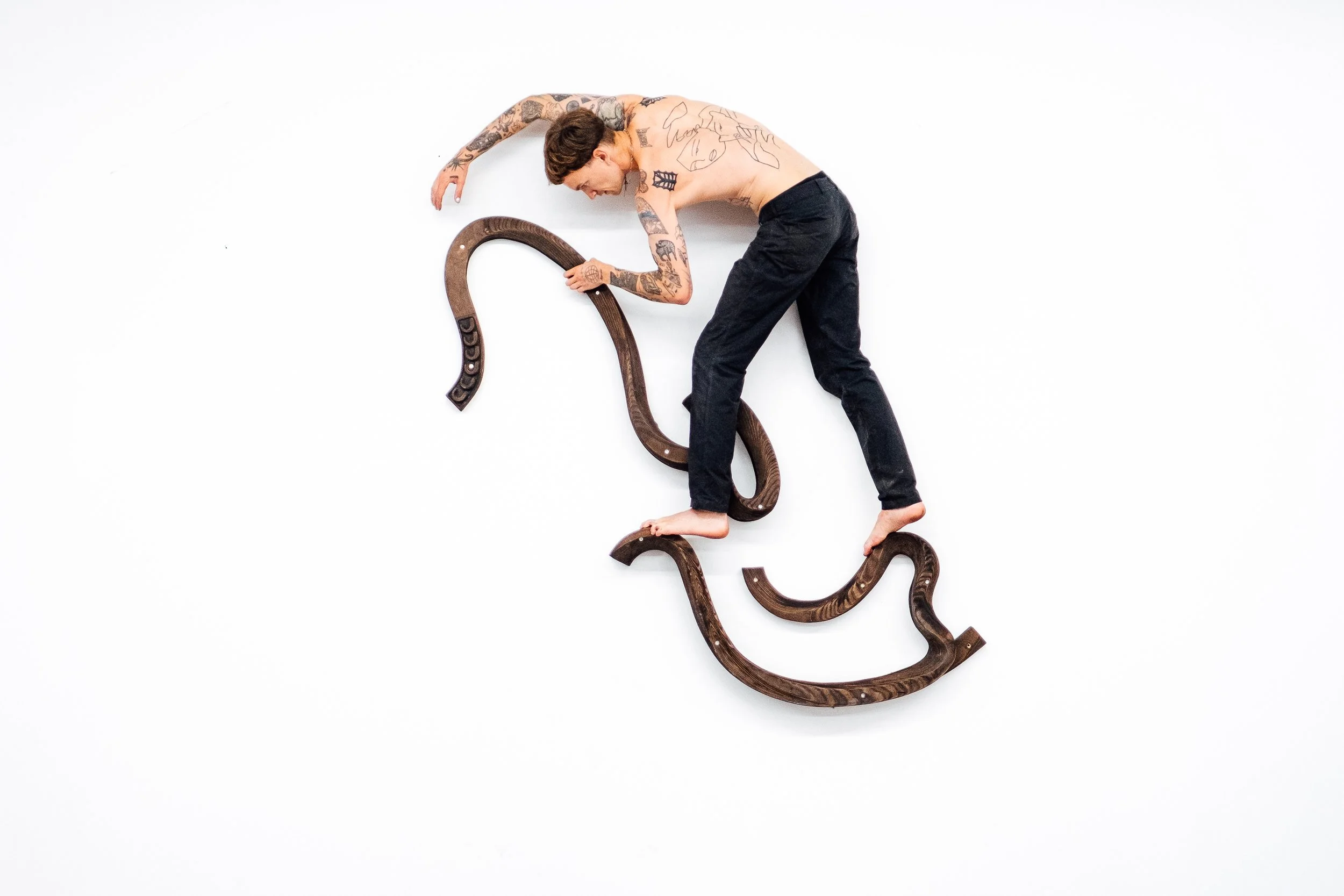 Un homme sans vêtements couché sur un mur blanc, tenant une pièce de bois courbée, avec de nombreux tatouages sur le corps.