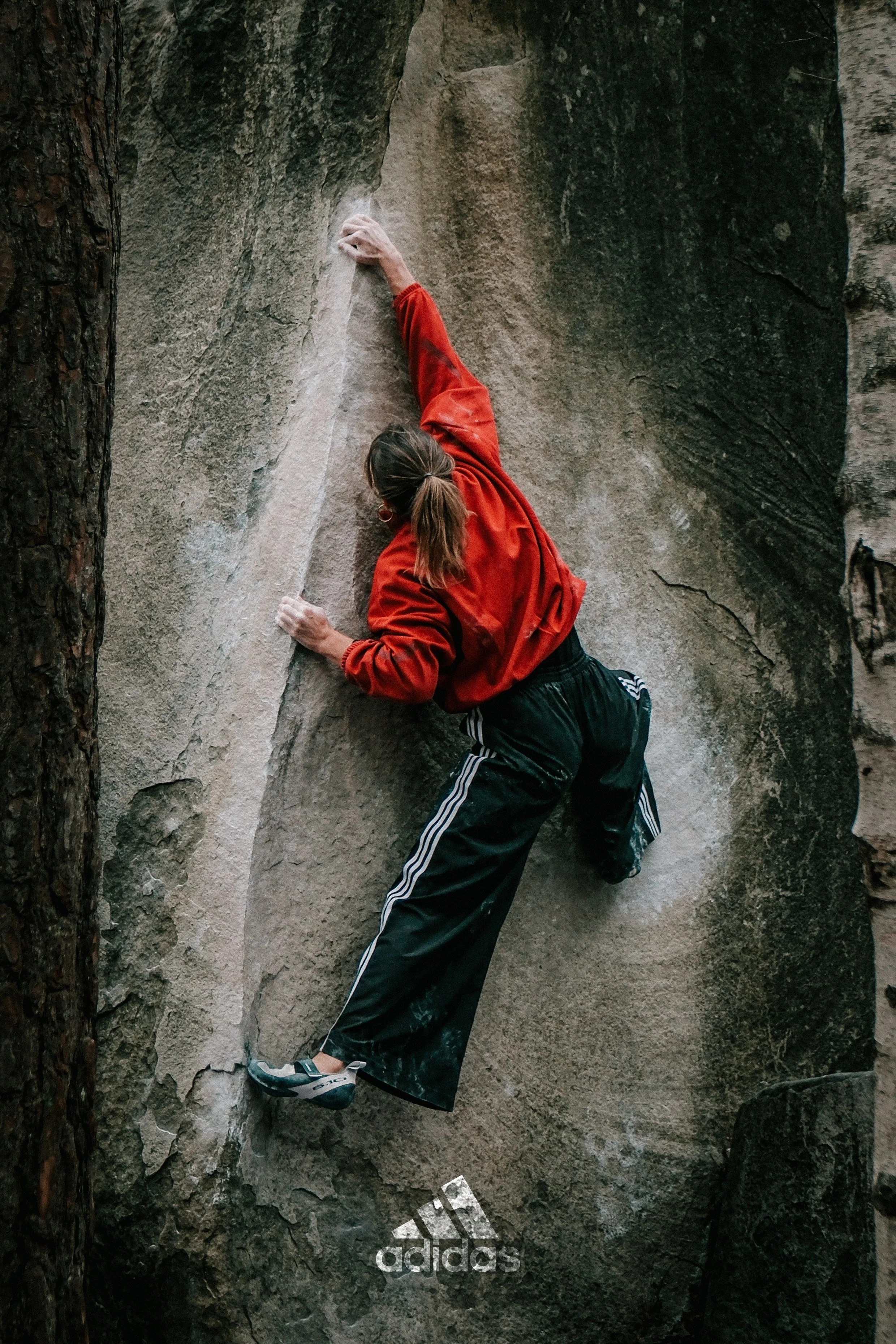 Une femme fait de l'escalade sur une paroi rocheuse, portant un pantalon noir, un sweat rouge, des chaussures de sport, dans une forêt.
