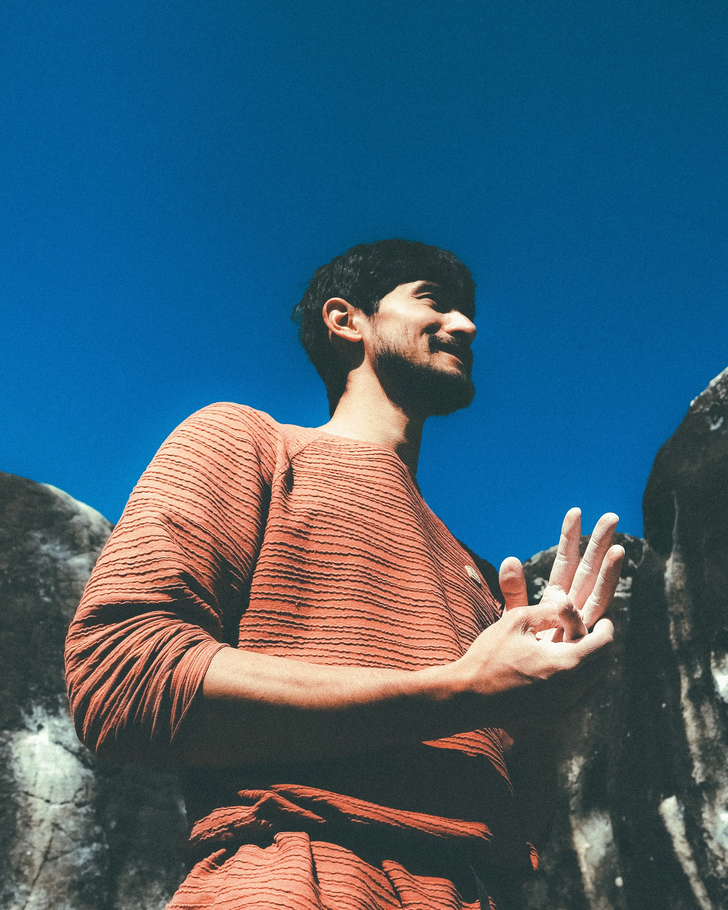 Jeune homme avec barbe coiffée, portant un pull rayé rouge et noir, levant la main dans un décor naturel avec rochers et ciel bleu.
