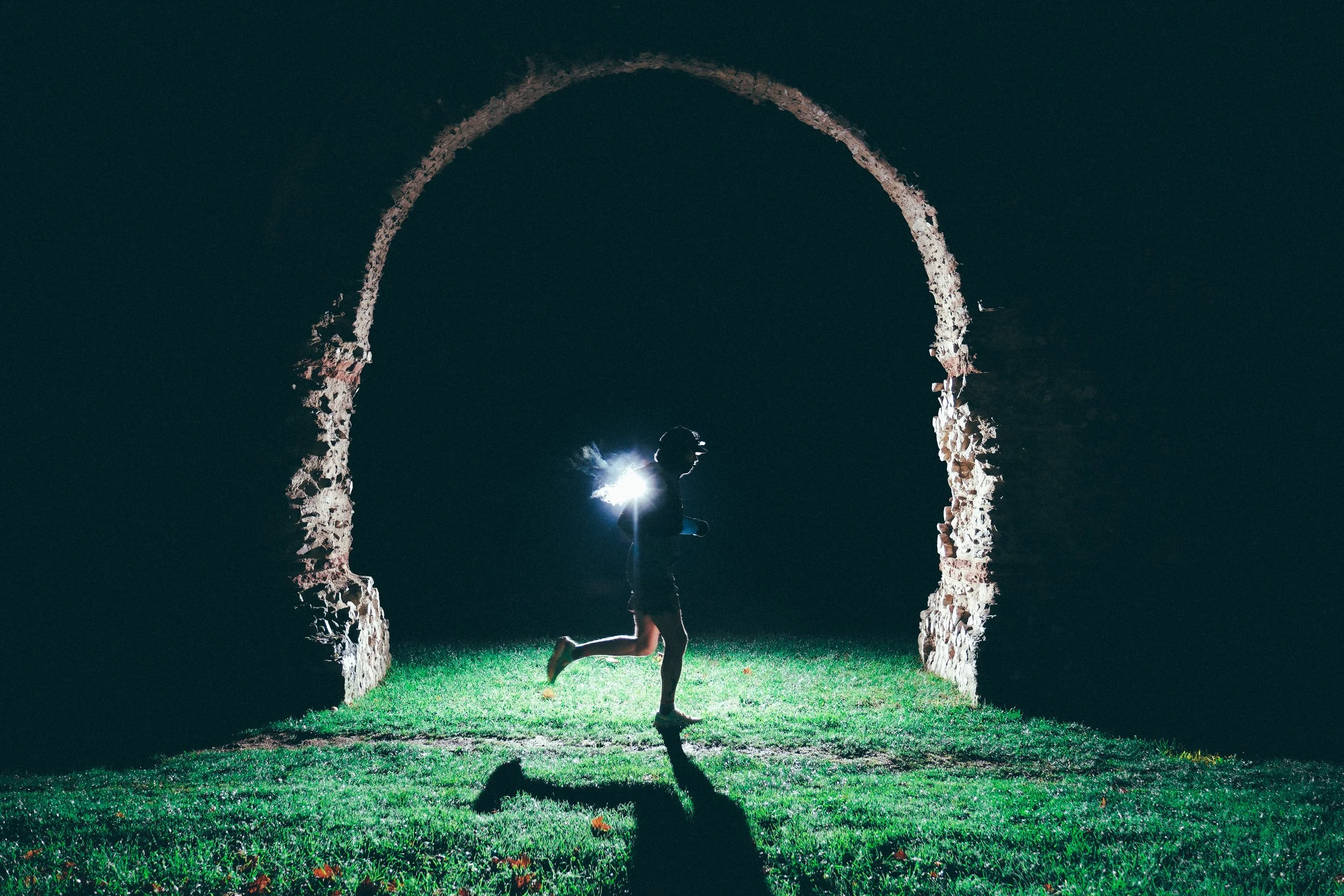 Une personne en train de courir dans une arche en pierre, éclairée par une lumière vive située derrière elle, la nuit.