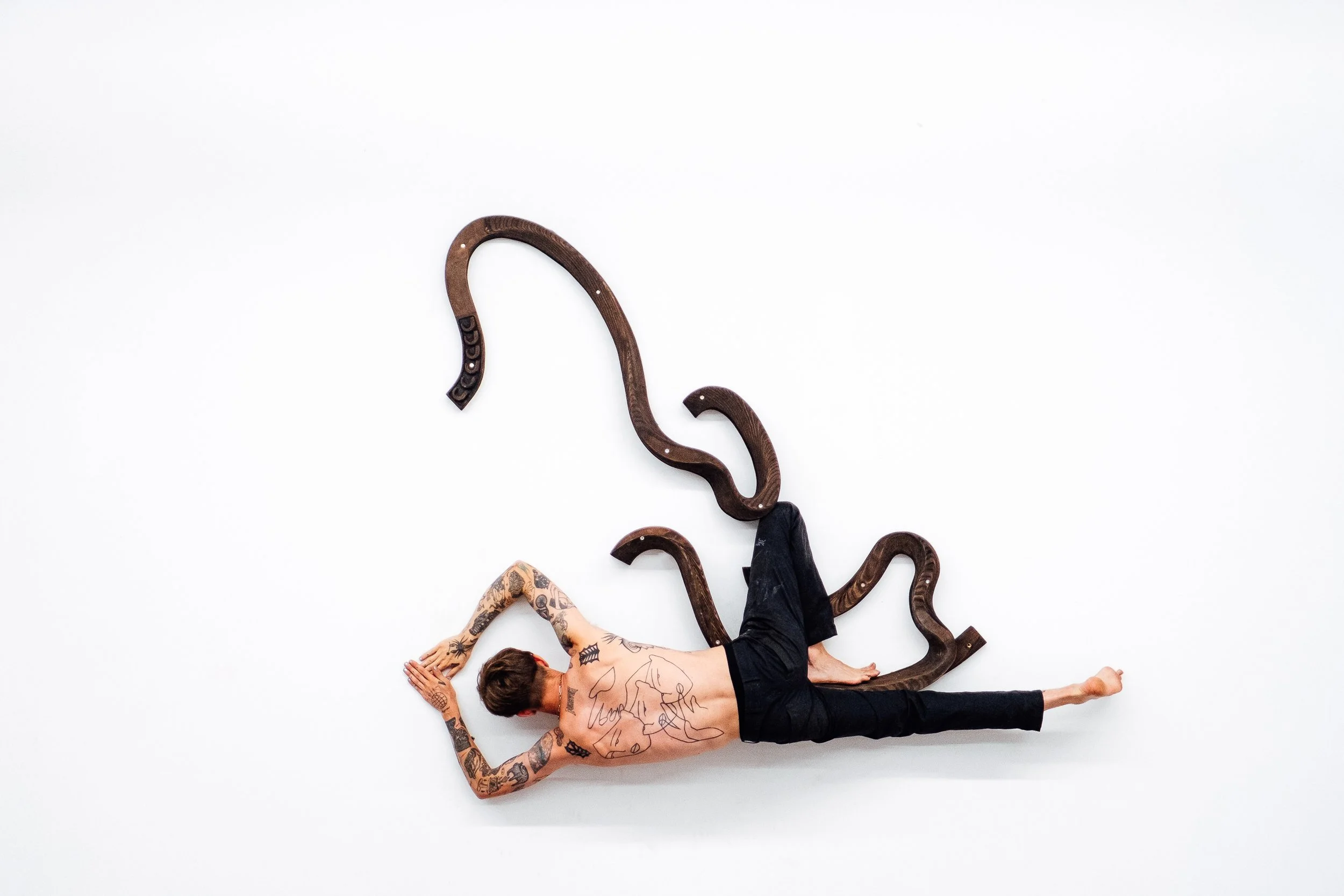 Un homme torsadé portant des tatouages, allongé sur le sol blanc, bras derrière la tête, avec plusieurs morceaux de bois en forme de serpents autour de lui.