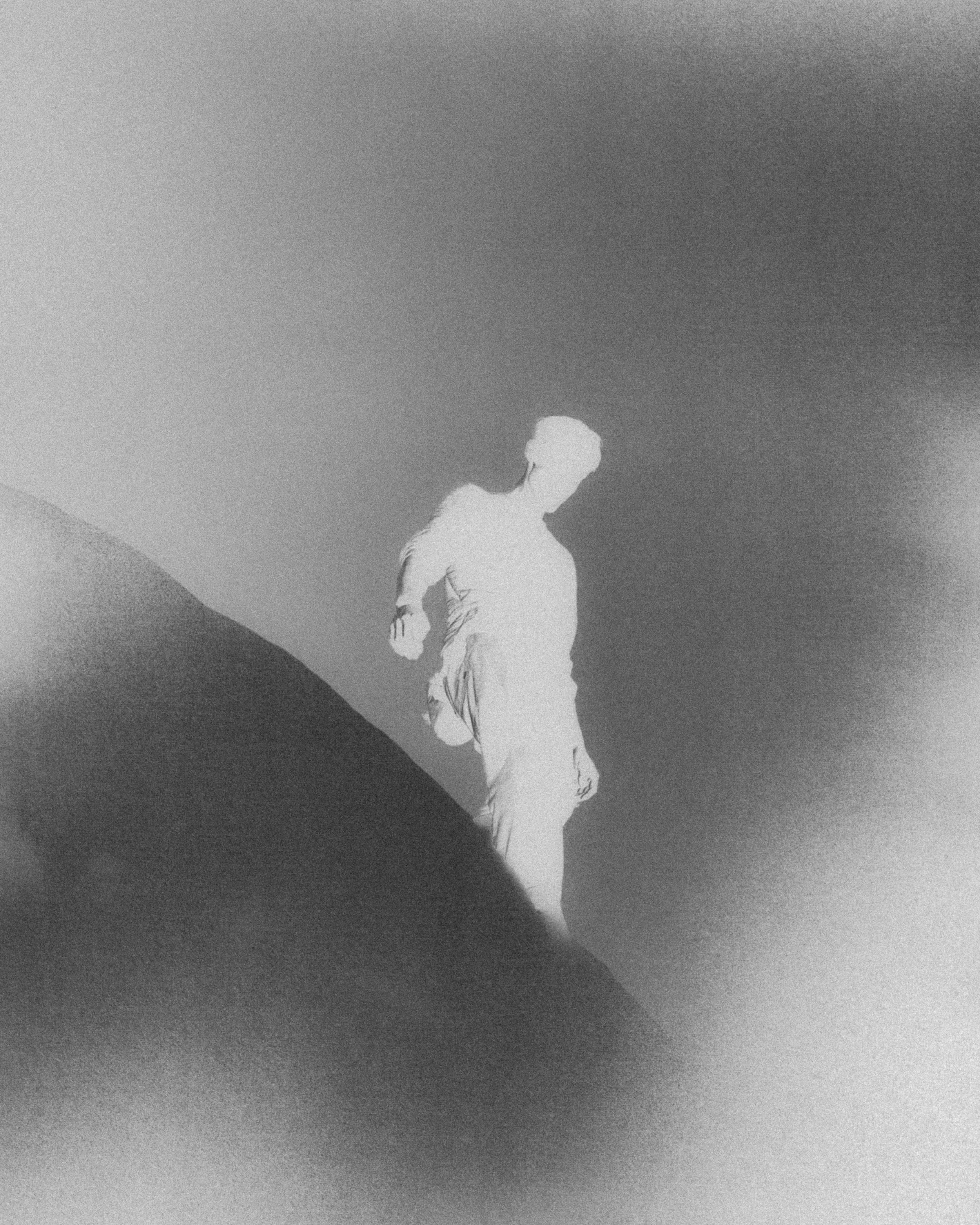 Silhouette d'une personne debout sur un terrain vallonné, regardant vers un ciel nuageux, en noir et blanc avec un effet de grain.