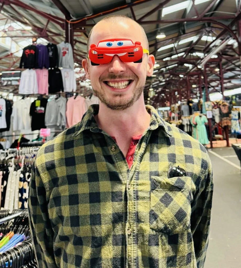 A man smiling at a market wearing a green and black checkered shirt and a pair of glasses in his shirt pocket. He is also wearing a red foam mask with cartoon eyes and a racing car design over his eyes, in an outdoor shopping area with various clothing items hanging on racks in the background.