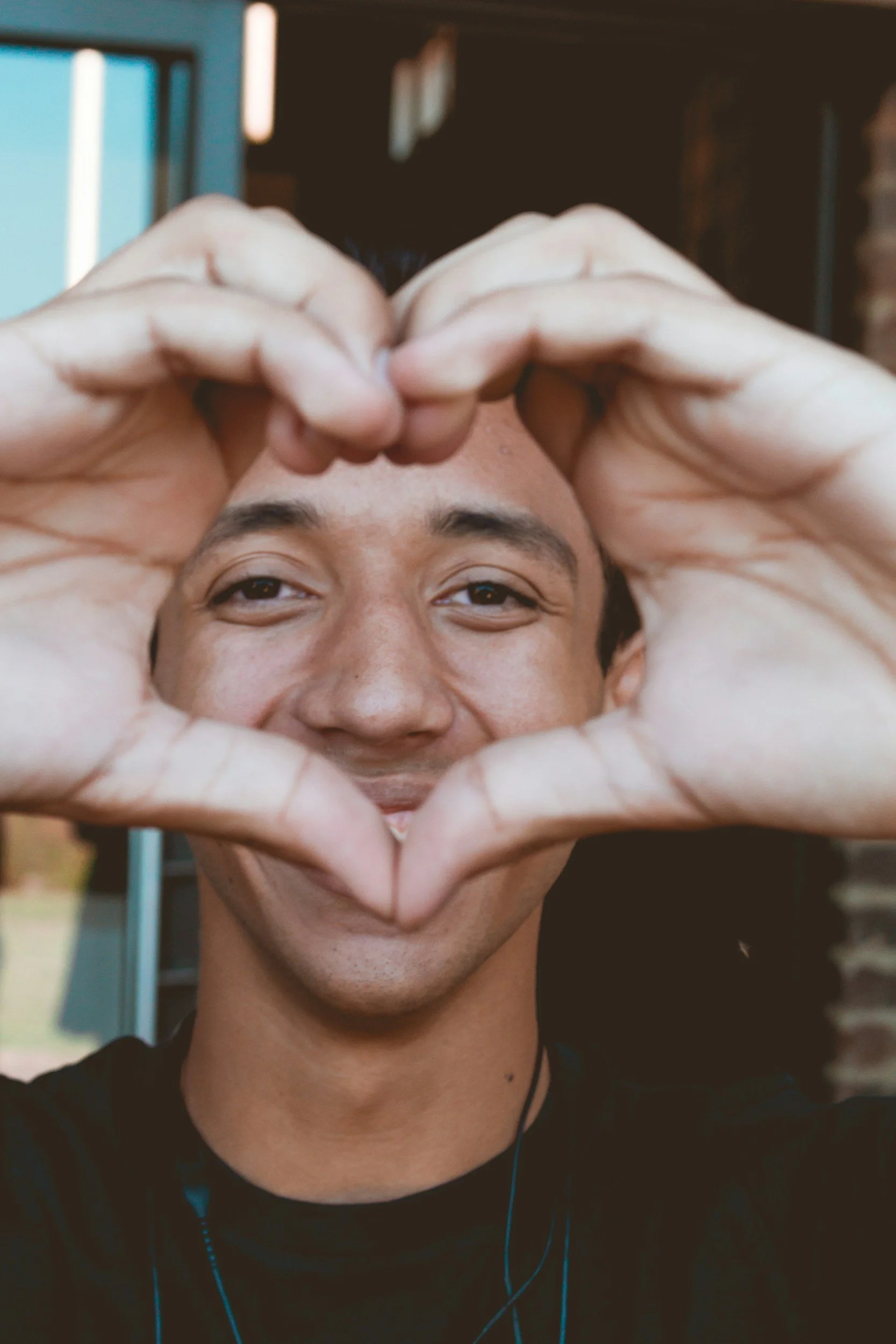 A young man smiling at the camera, holding his hands up in a heart shape around his face. He is wearing headphones and a black shirt, with a blurred indoor background.