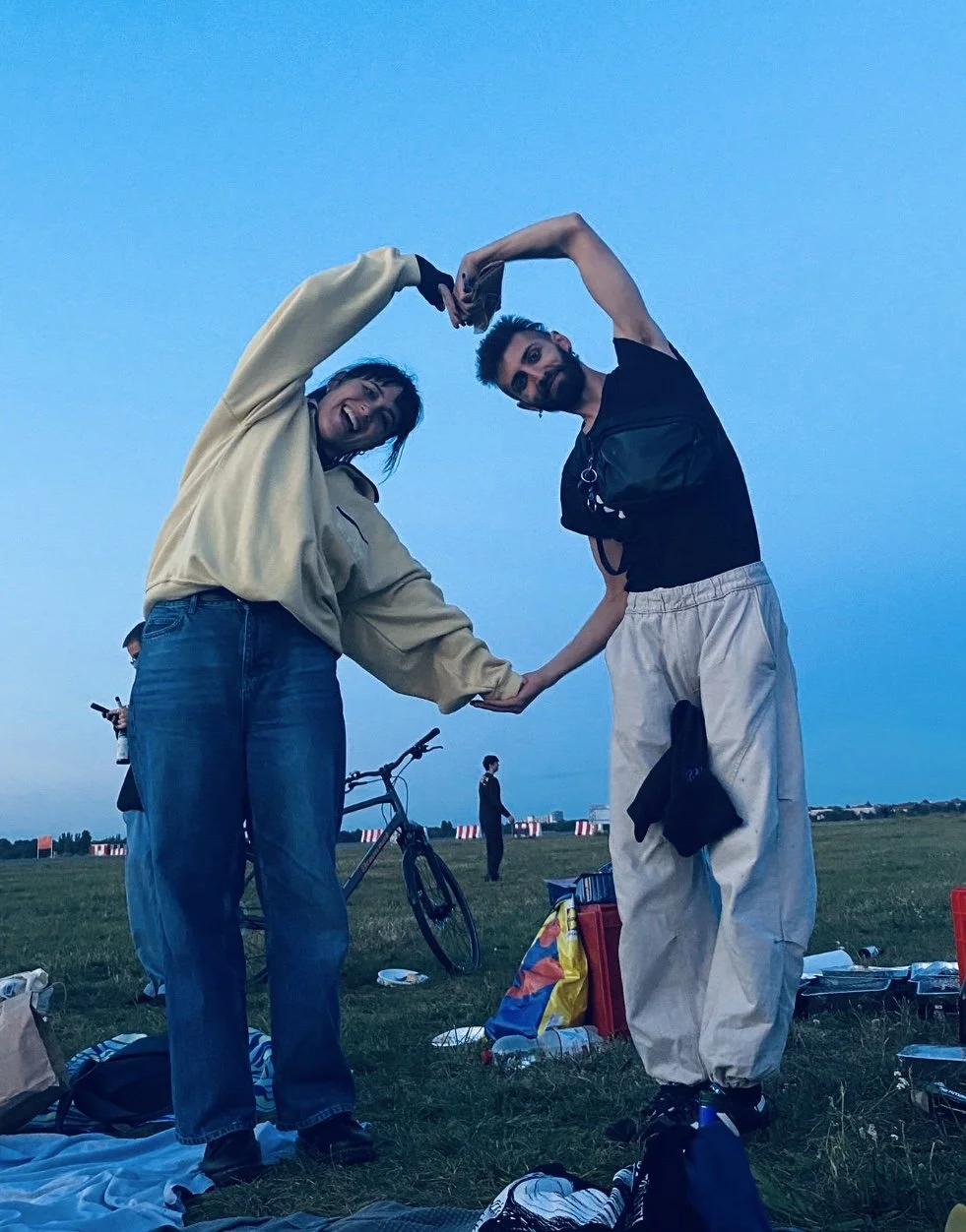 Two people are forming a heart shape with their arms and holding hands, smiling at the camera. They are outdoors in a grassy area during dusk, with other people, a bicycle, and picnic items visible in the background.
