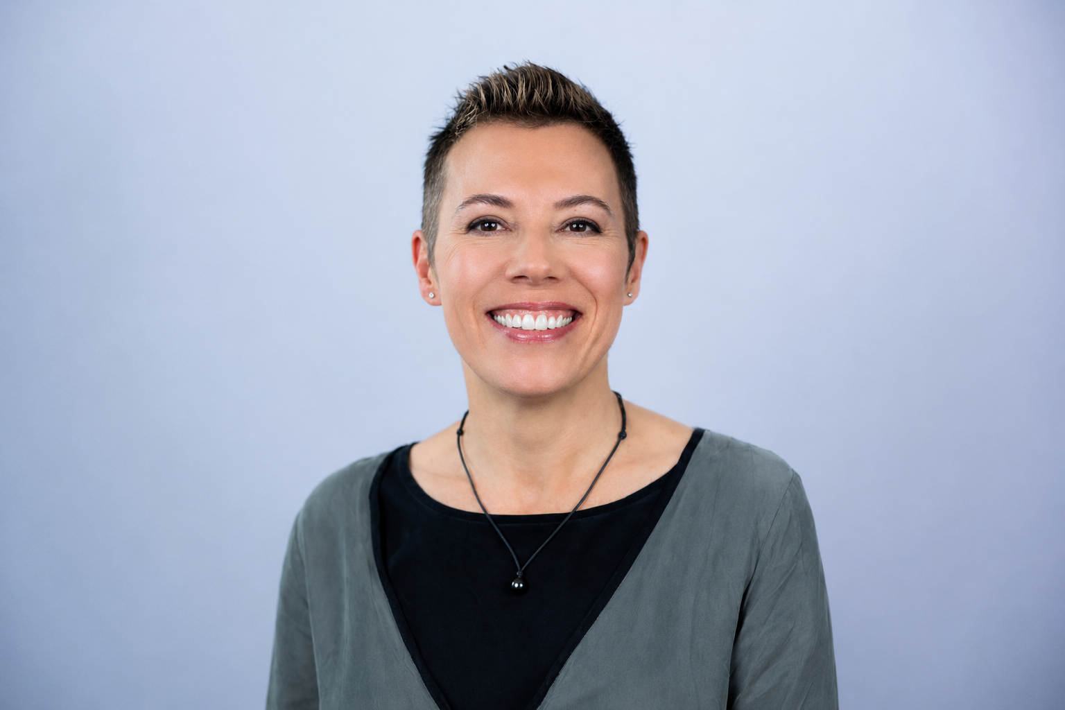 A smiling woman with short dark hair, wearing a black top, grey jacket, and a black beaded necklace, standing against a plain light blue background.