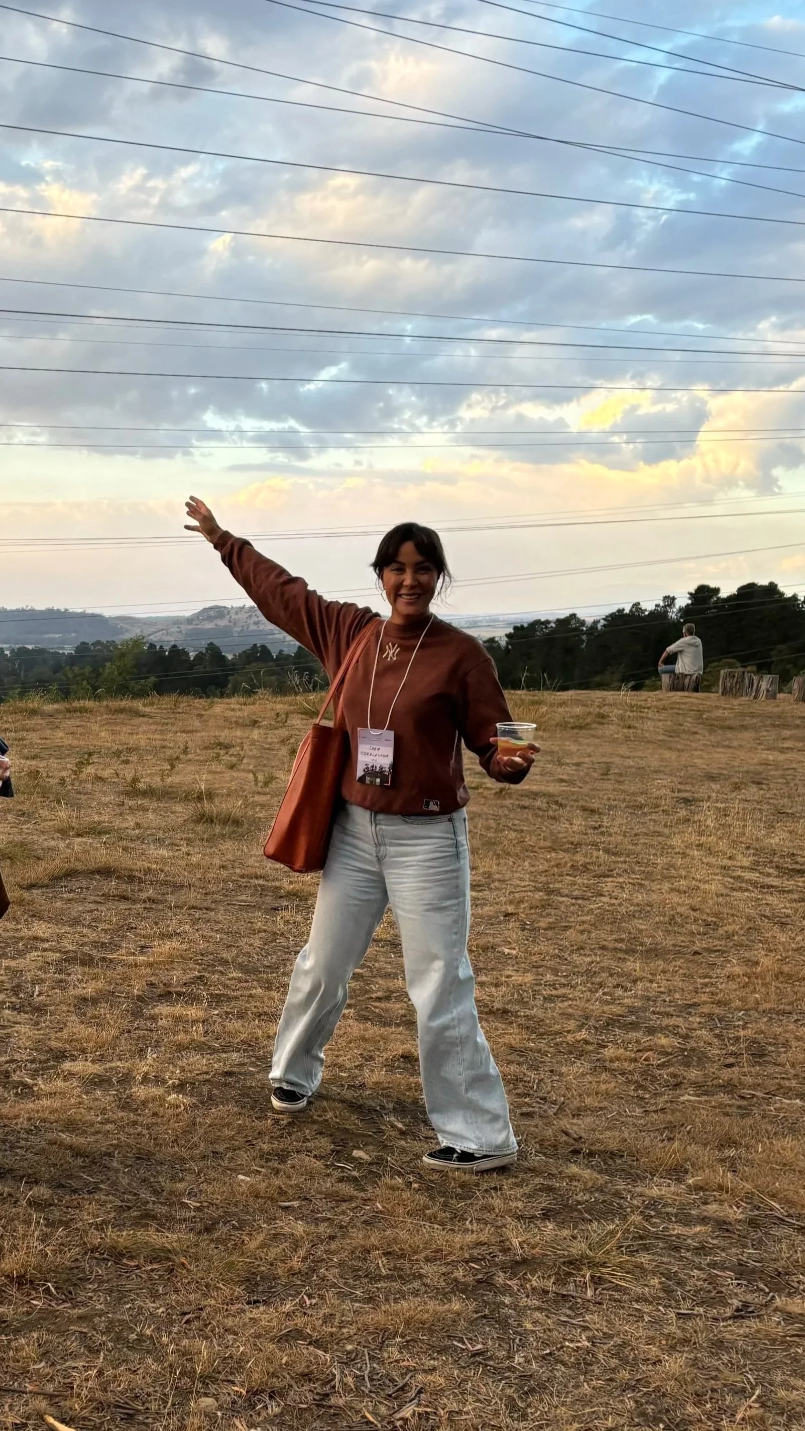 A woman standing outdoors on dry grass, smiling and gesturing with her right arm raised, holding a drink in her left hand, with power lines and cloudy sky in the background.