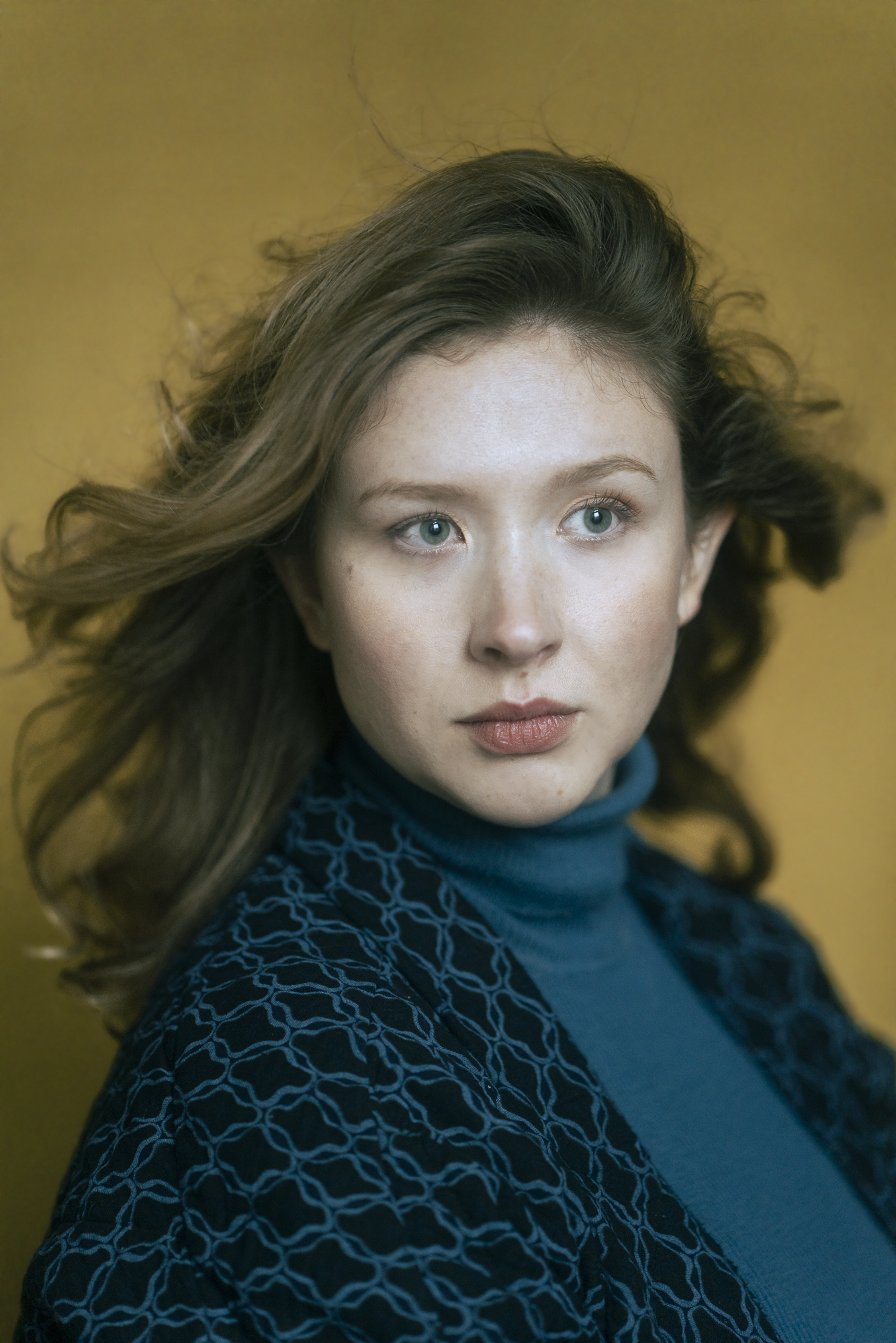 Portret van een jonge vrouw met lang, golvend haar en blauwe ogen, gekleed in een donkerblauw patroonhemd, tegen een gele achtergrond.