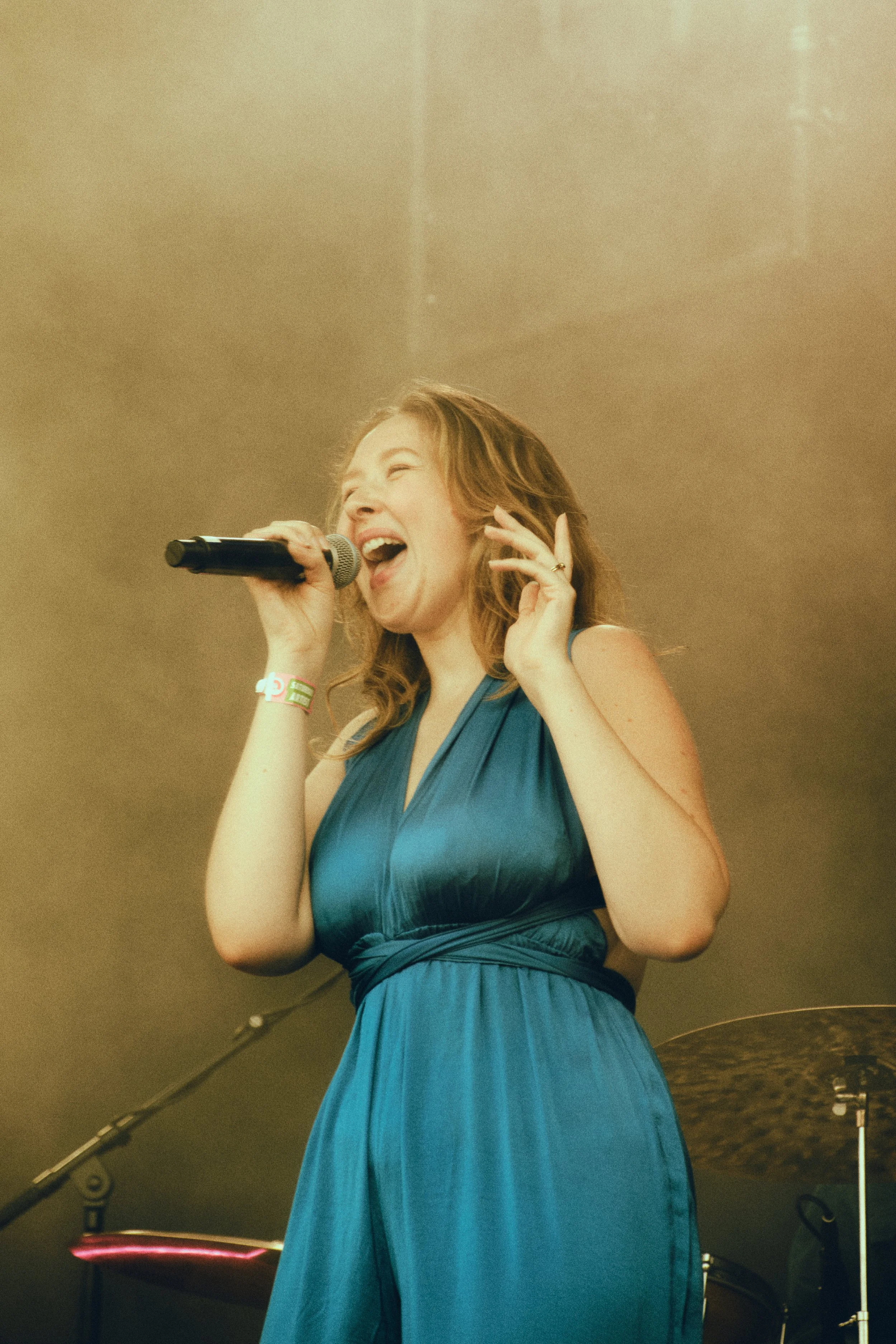 Vrouw zingt in een blauw jurk met microfoon, lachend, in een lichte zaal.