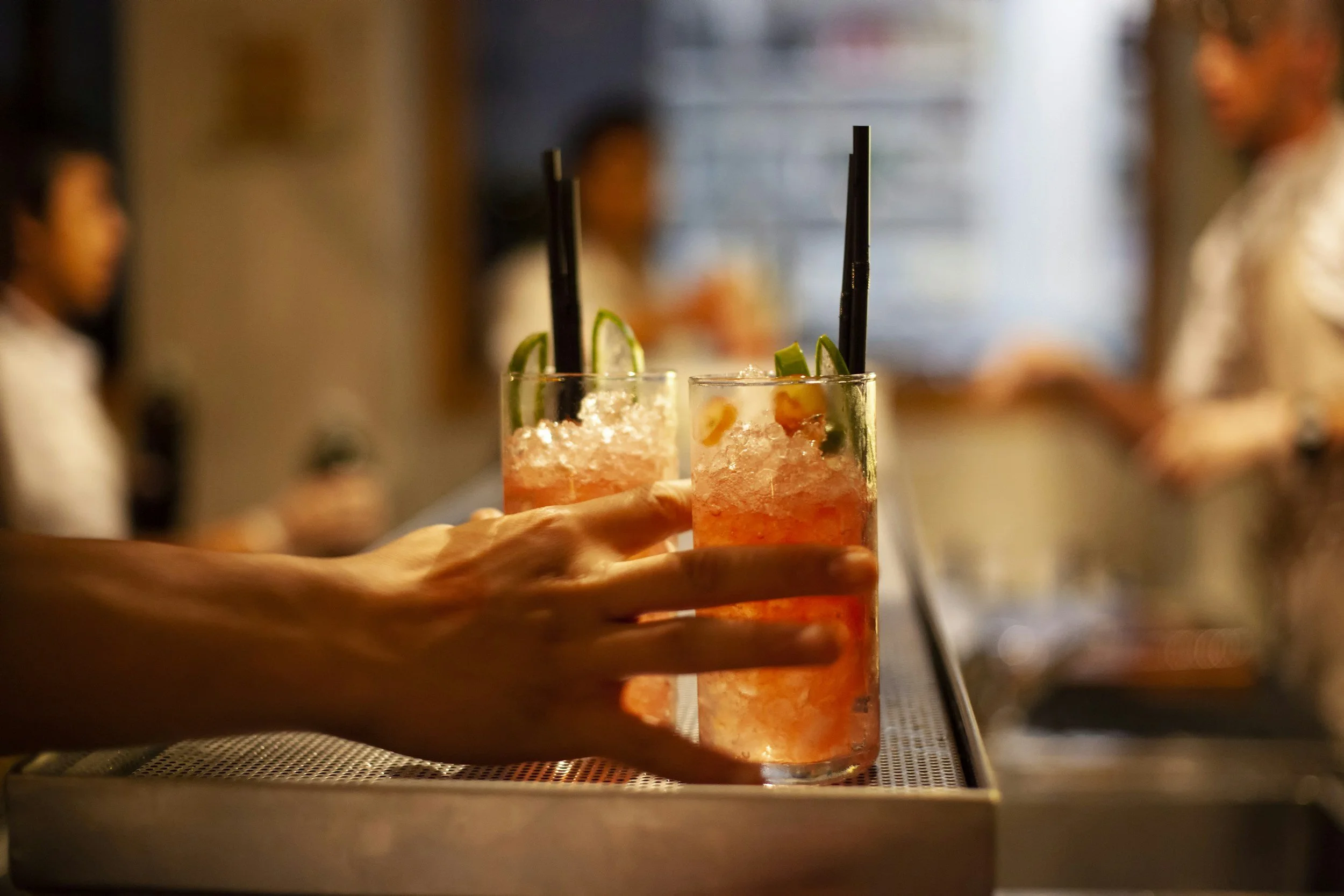 Two colorful cocktails with crushed ice, garnished with lime slices, and black straws, being served at a social event with blurred people in the background.
