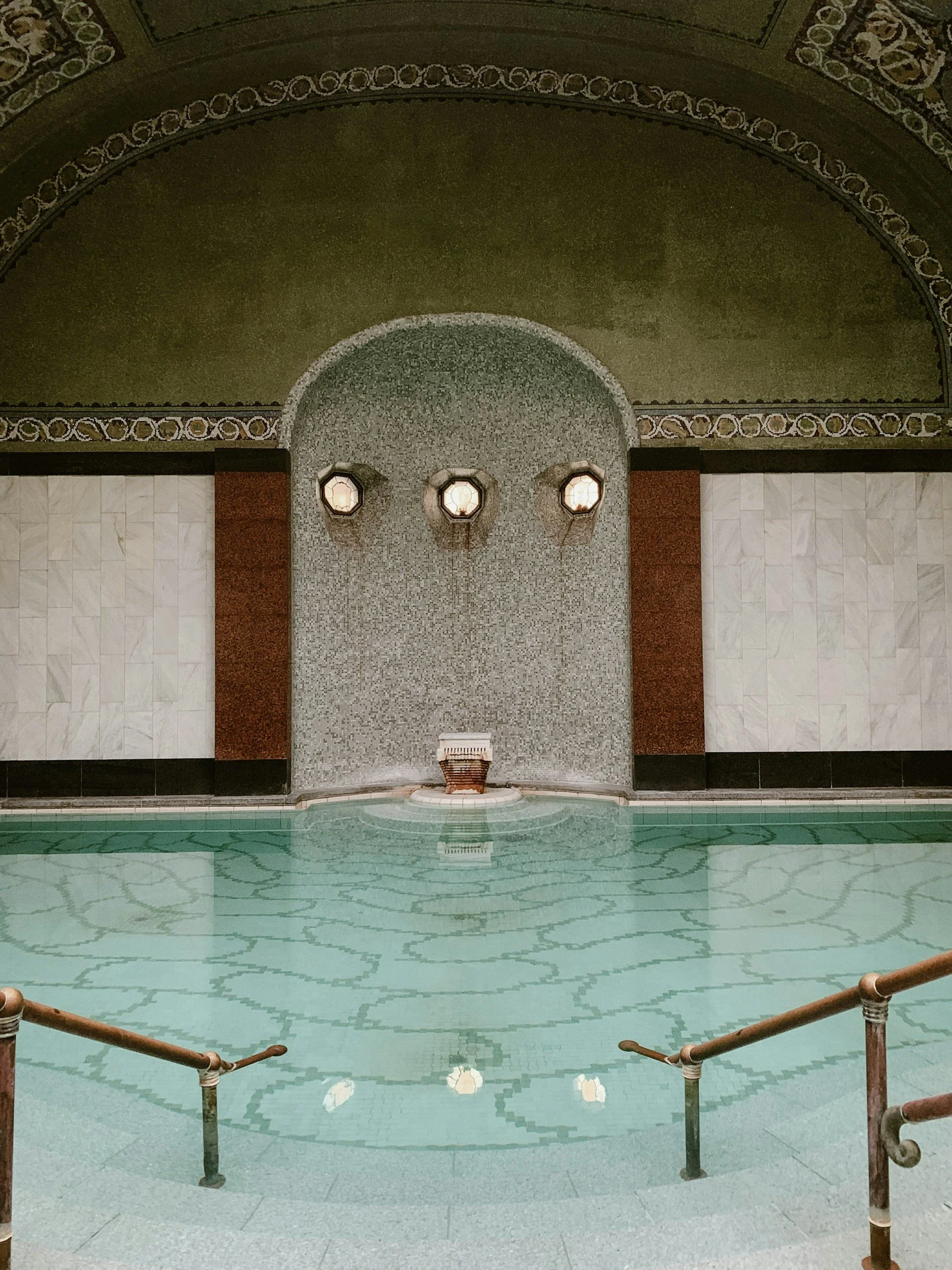 Interior view of a luxury swimming pool with ornate walls and arches, featuring three hanging lamps on a mosaic wall, surrounded by decorative railing and marble flooring.