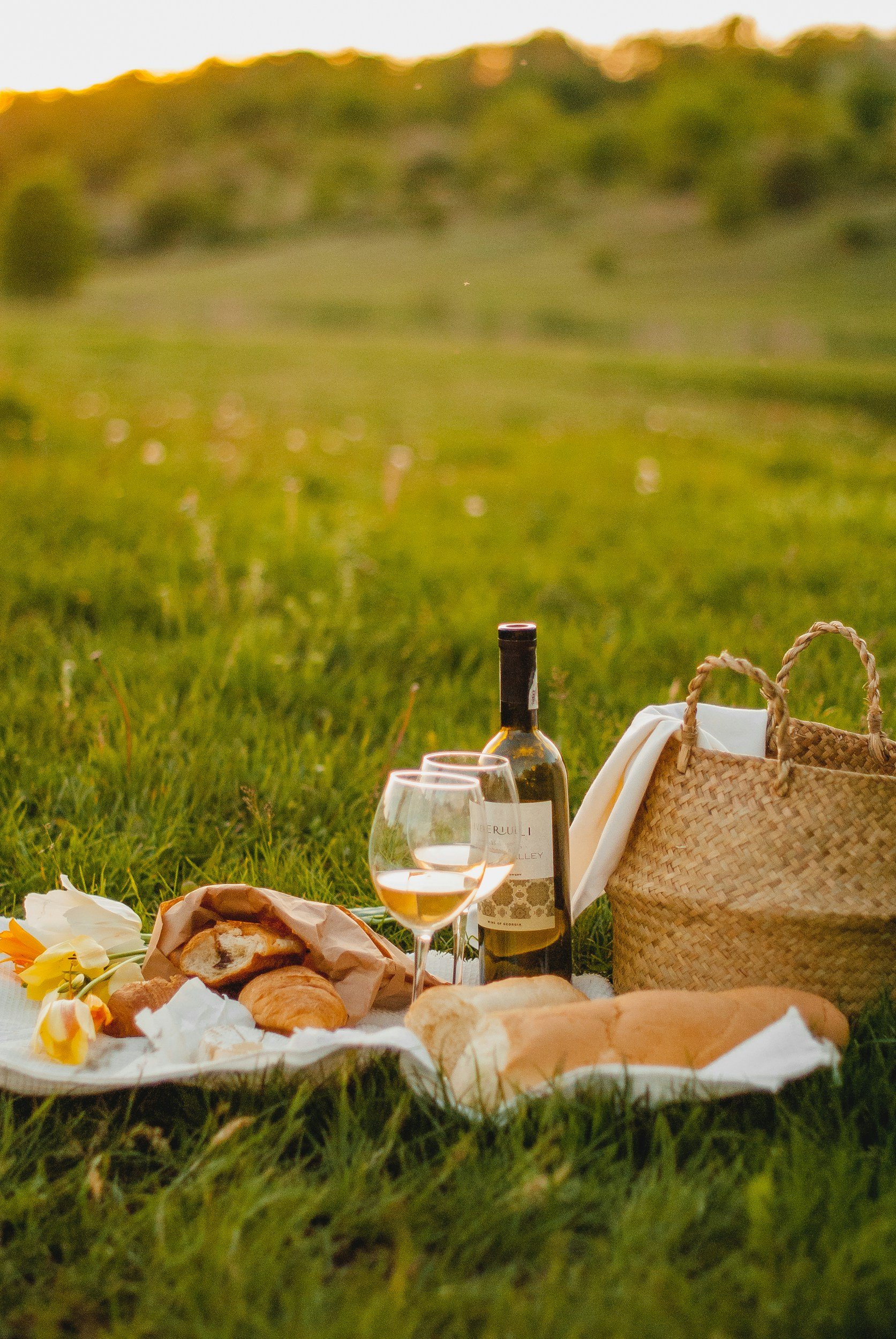 Picnic setup on grass with wine, glasses, bread, pastries, flowers, and a woven basket during sunset in a rural setting.