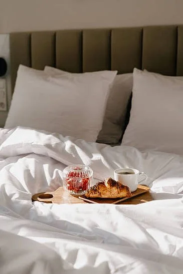 A bedroom with a bed that has white pillows and bedding. On the bed, there is a wooden tray with a croissant, a cup of coffee, and a jar of jam.