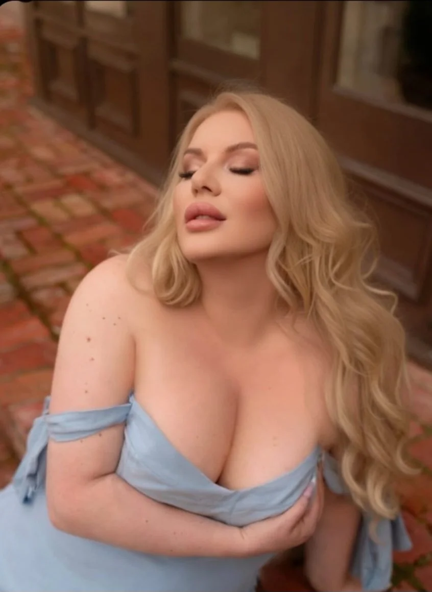 A woman with long blonde hair, closed eyes, and nude makeup, wearing an off-the-shoulder light blue dress, posing outdoors on a cobblestone street.