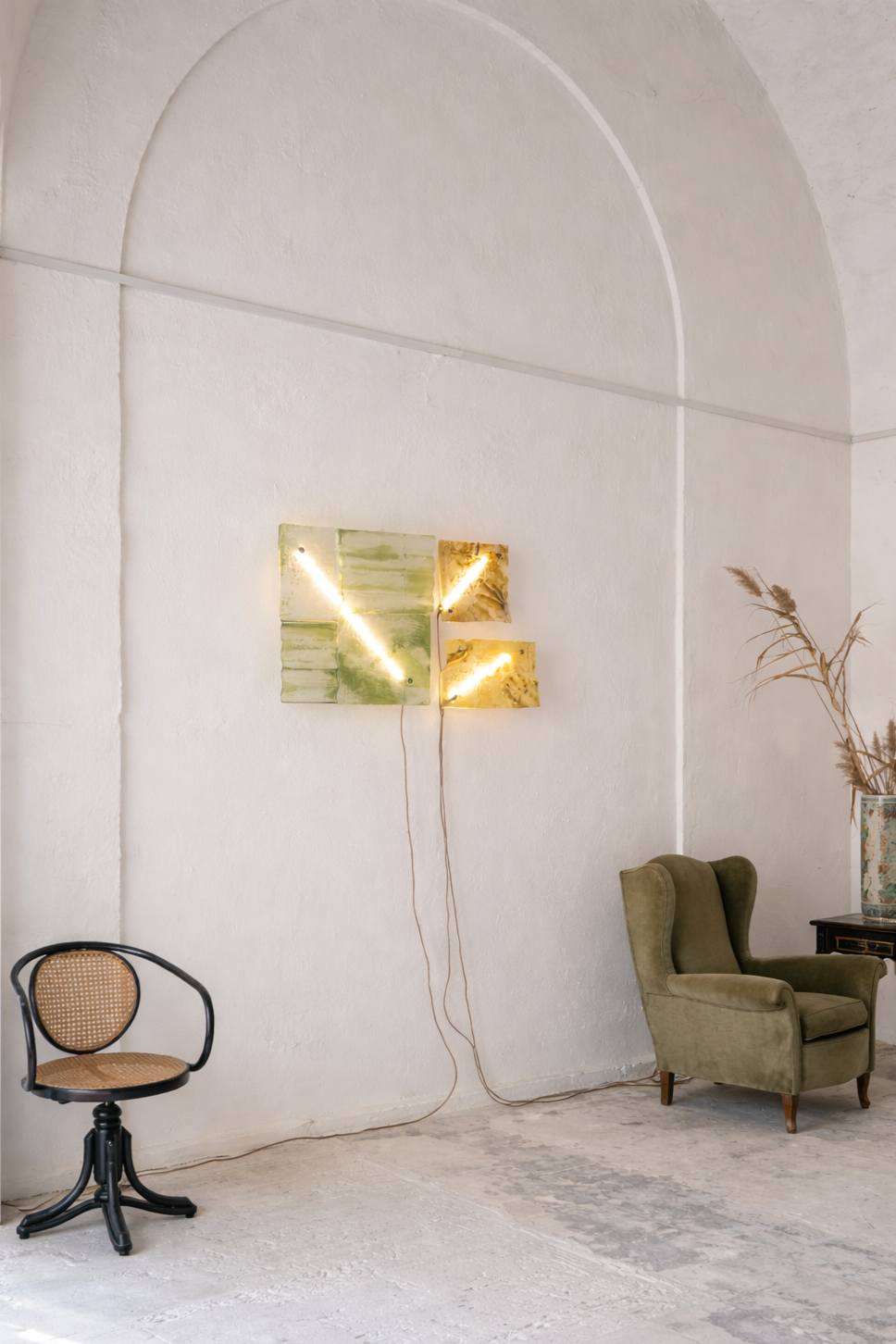 Interno con parete bianca, poltrona verde, sedia di paglia con struttura nera, decorazione con luci e foglie di palma secche in vaso.