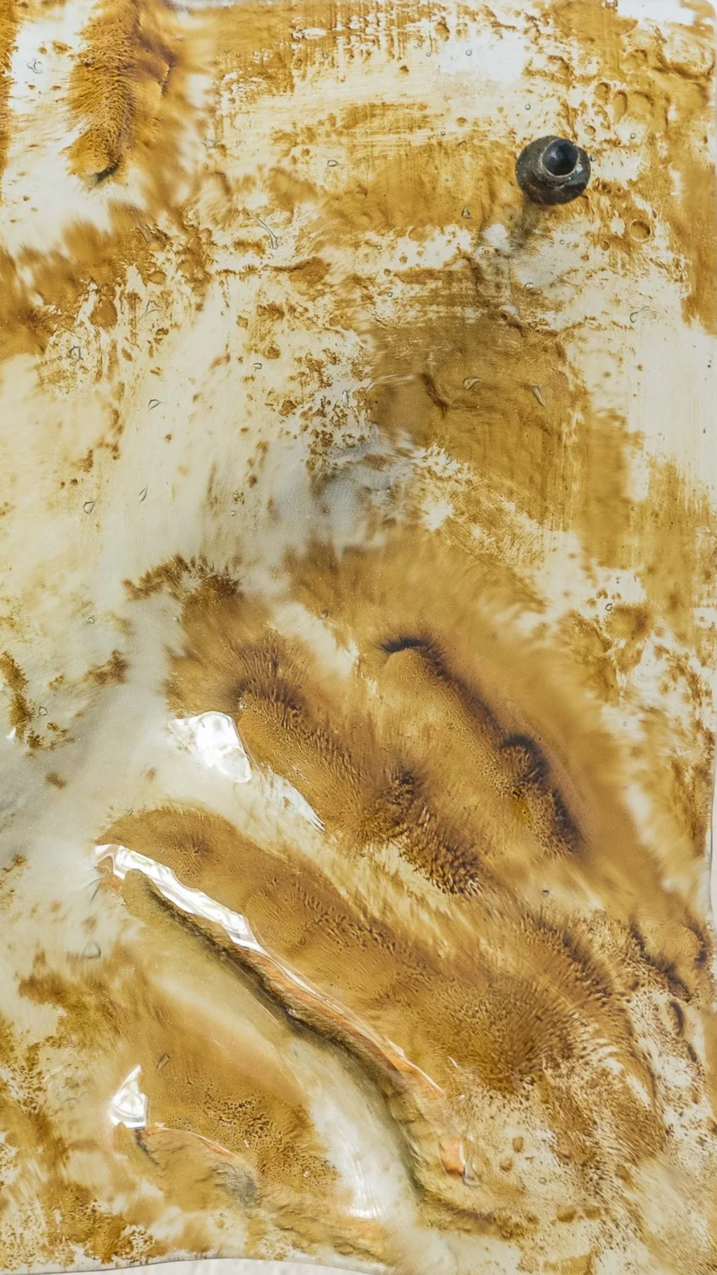 Superficie in vetro con tracce di colore marrone e crema, con alcune macchie e una vite nera nell'angolo superiore destro.