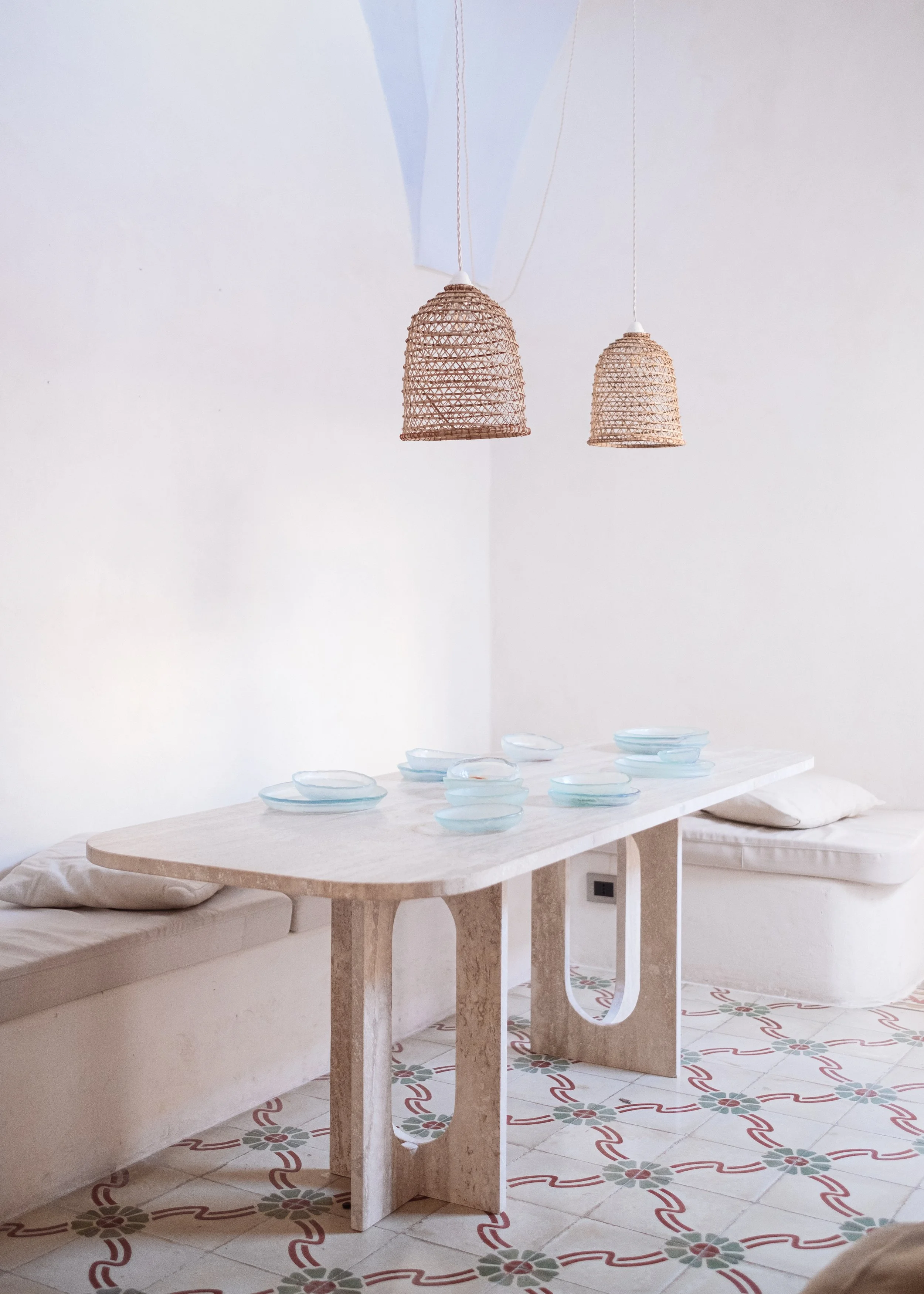 Interno di una stanza con un tavolo di pietra con una seduta imbottita, decorata con piatti di ceramica azzurri sopra un tavolo, due lampade di paglia sospese dal soffitto, pavimento con piastrelle decorative con motivi floreali e linee rosse.