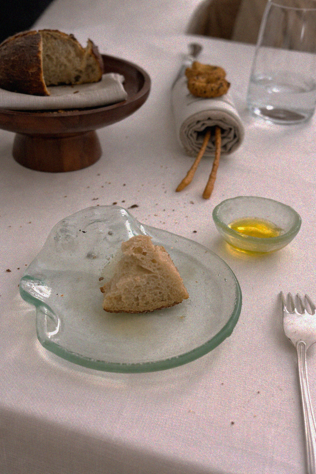 Particolare di una tavola imbandita con un pezzo di pane su un piattino di vetro trasparente, una ciotola di olio, una fetta di pane, alcuni biscotti, un bicchiere d'acqua, e una rotella di pane con degli stuzzicadenti sopra.