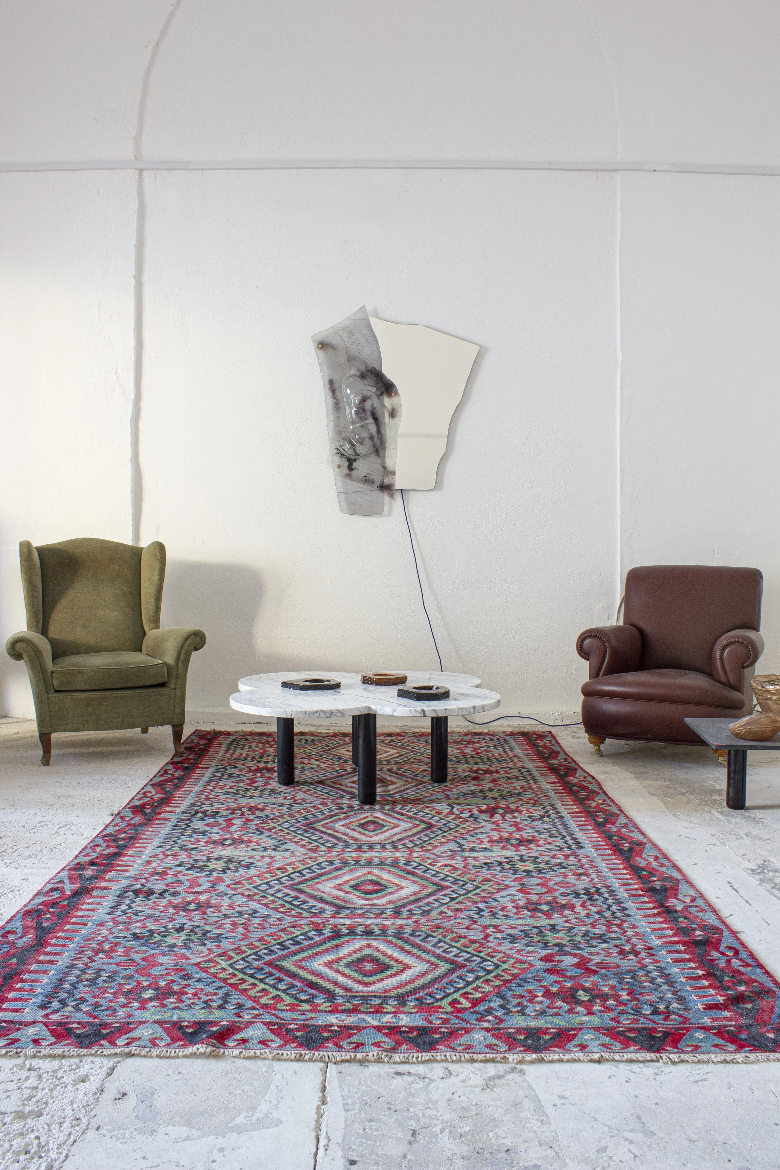 Interno con due poltrone, uno verde e uno rossa, un tavolino centrale con riviste e un tappeto persiano, pareti bianche con una scultura artistica