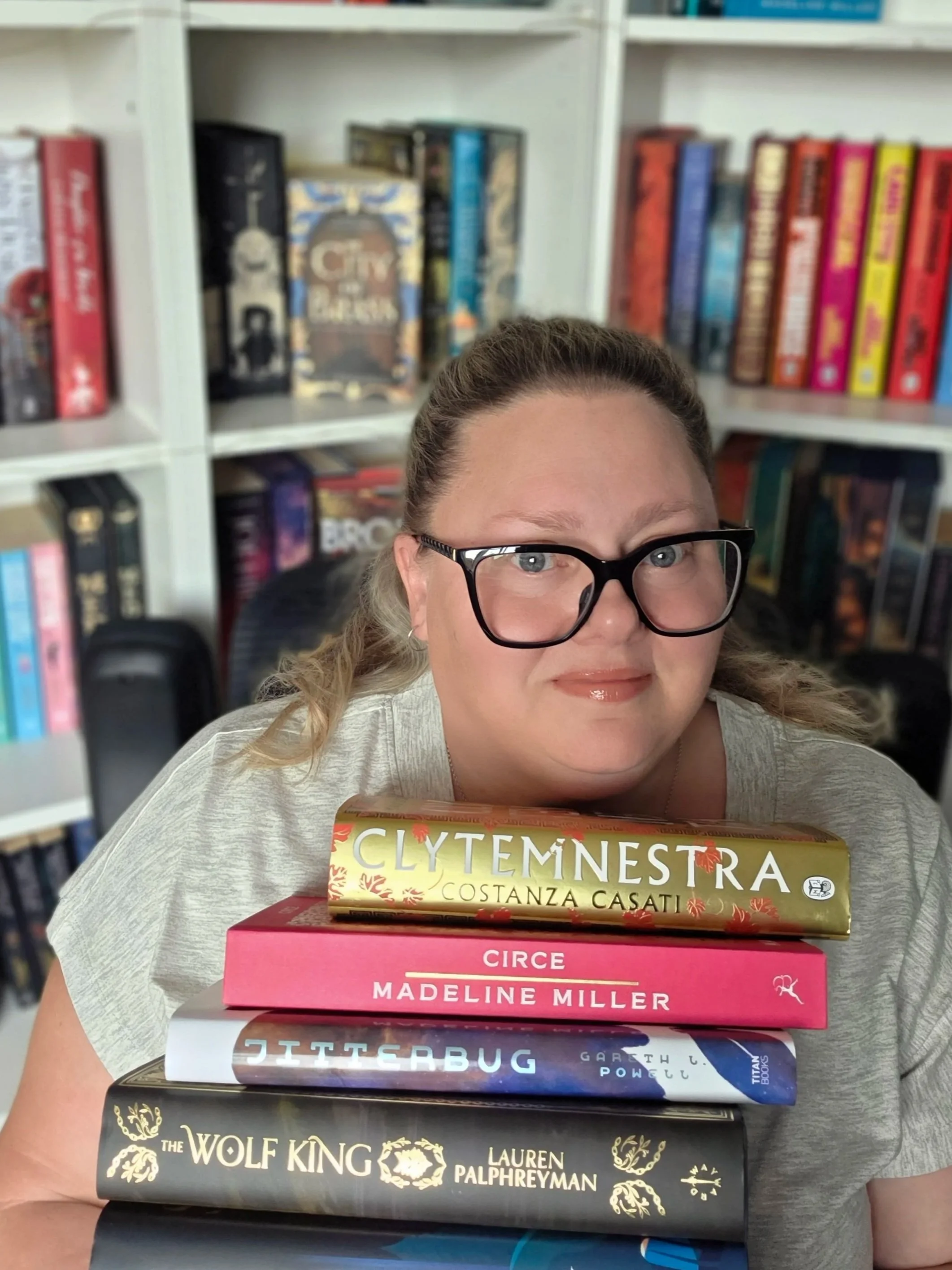 A woman with glasses and curly hair sitting in front of a bookshelf with a stack of books in her lap. The books include 'Clytemnestra' by Costanza Casati, 'Madeline Miller' by Circe, 'Gitterbug' by Gareth, and 'The Wolf King' by Lauren Palphreyman.