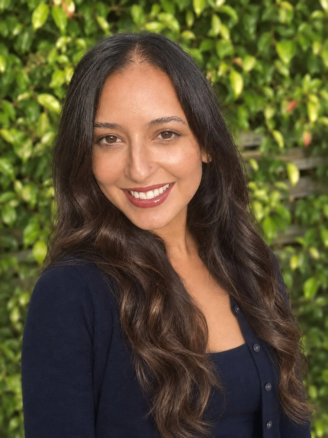 Smiling woman with long dark hair posing outdoors in front of green foliage